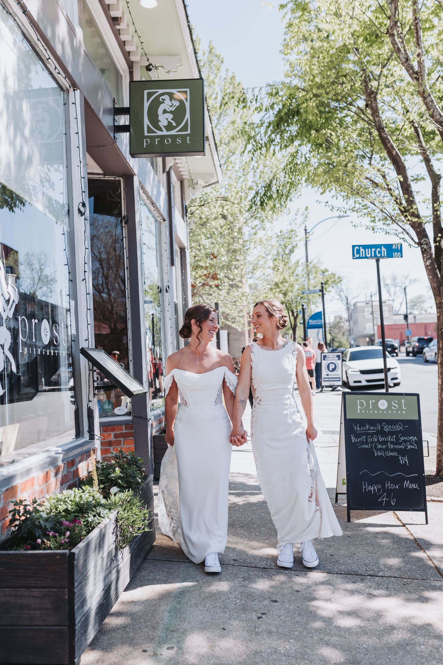 Two brides in white dresses hold hands while strolling past a boutique on Church Street.