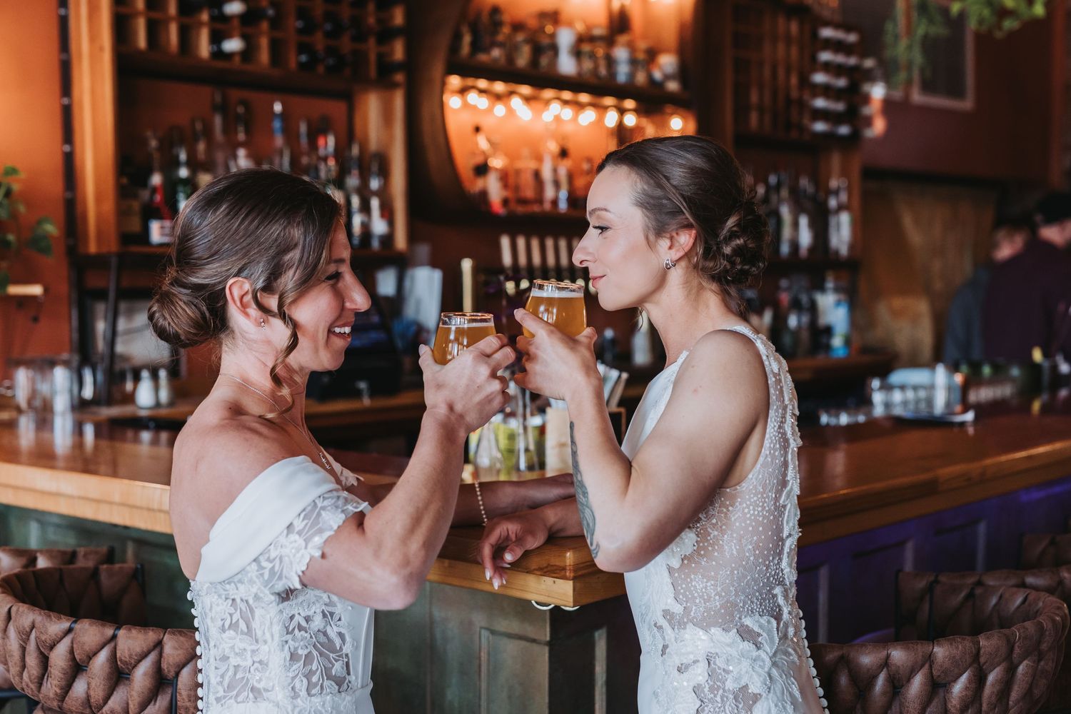 Two brides in white lace gowns toast with cocktails at a cozy bar venue.