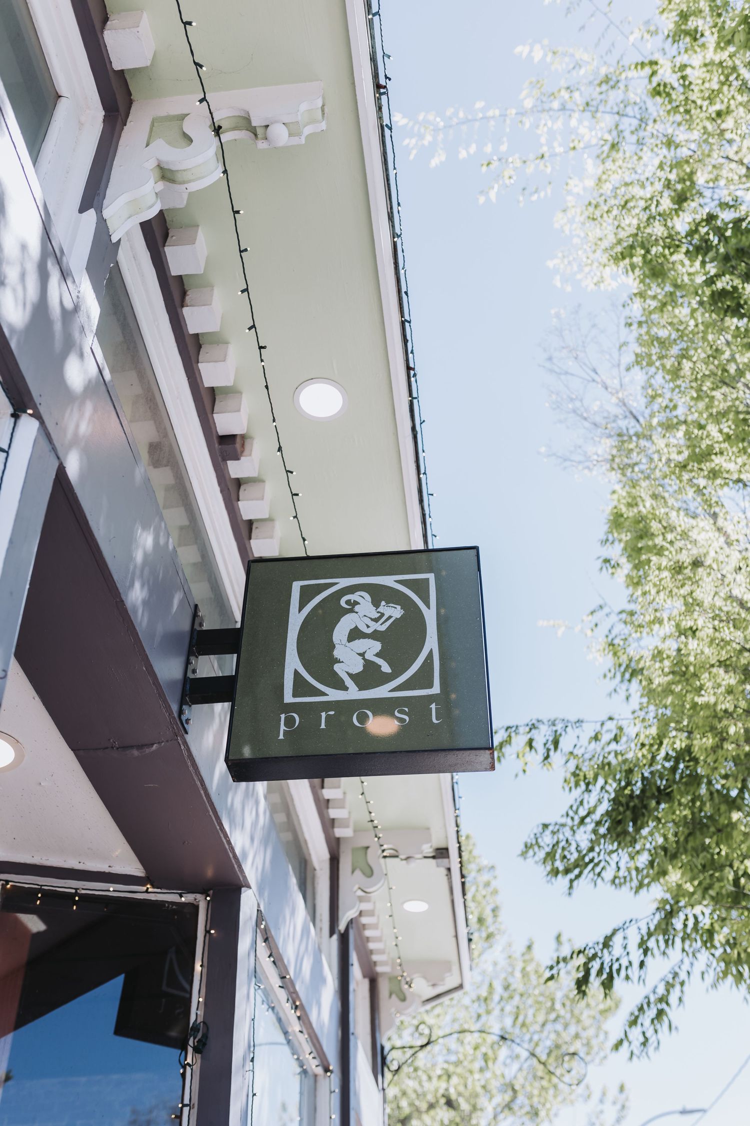 Exterior sign for Prost restaurant featuring a lion logo, hanging beneath a white building overhang with string lights.