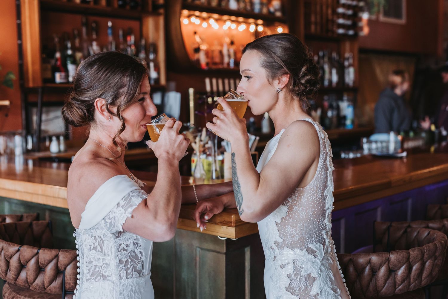 Two brides in white wedding gowns toast drinks together at a warmly lit bar.
