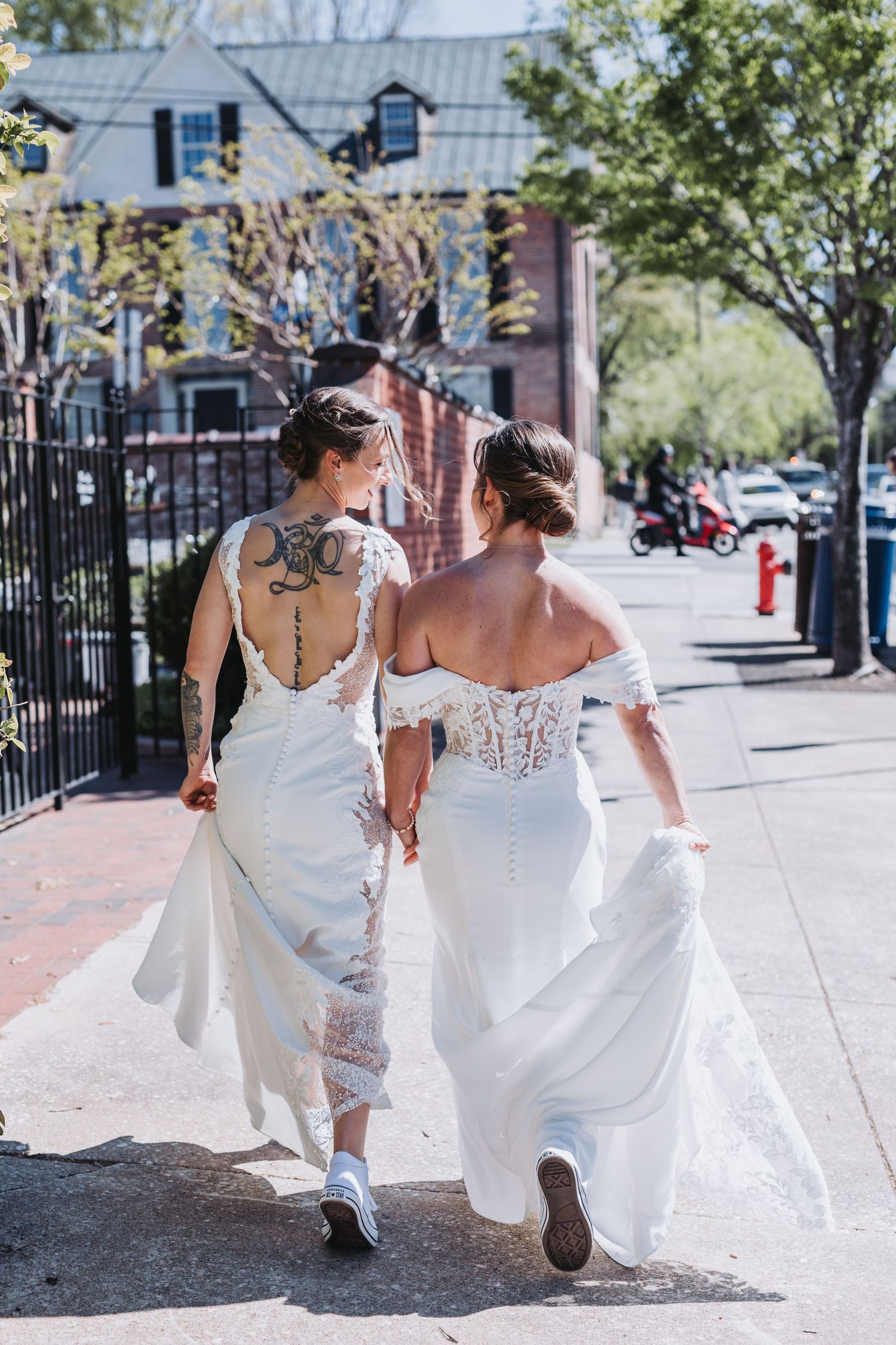 Two brides in white wedding dresses walk hand-in-hand down a sunny sidewalk, showing their backs.