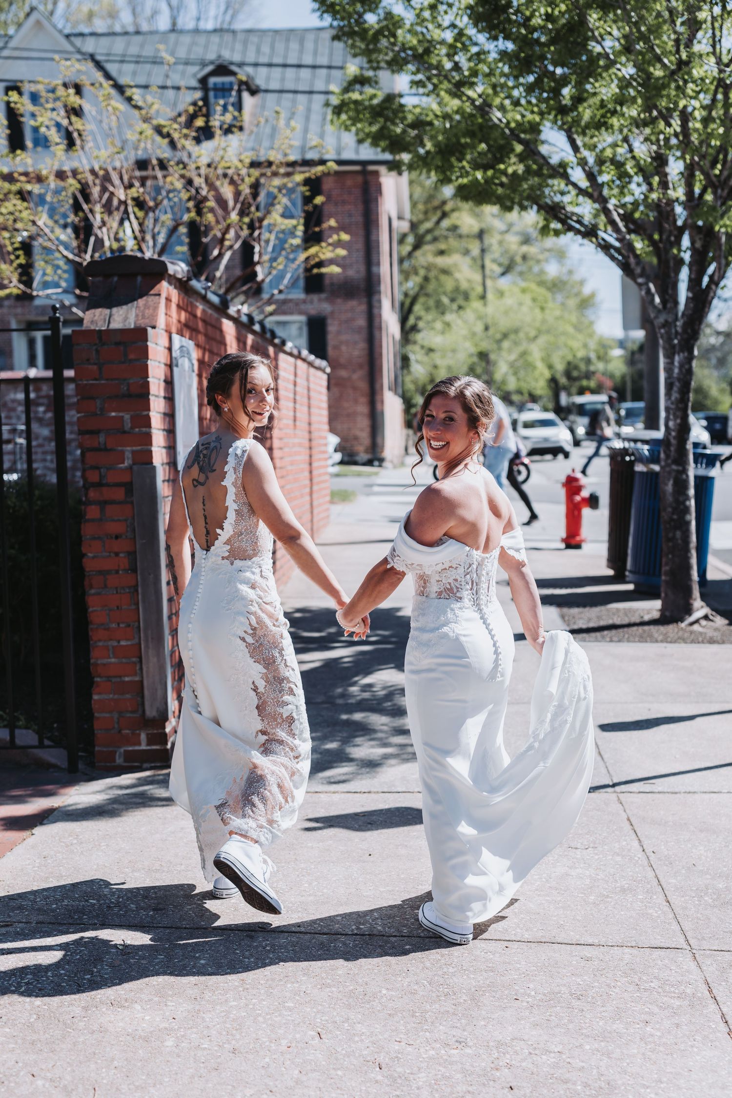 Two brides in white wedding dresses laugh and hold hands while walking down a sunny sidewalk.