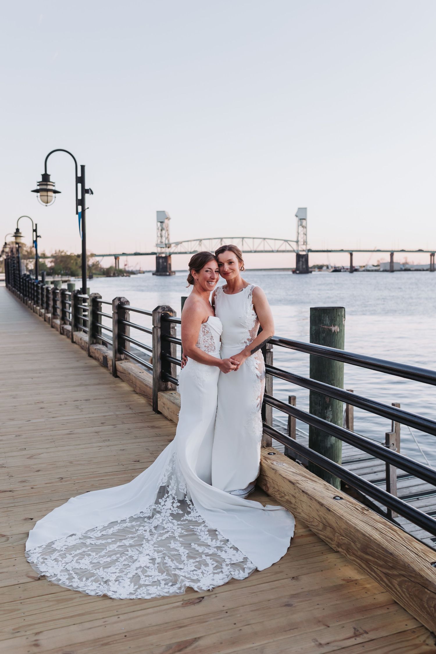 Two brides in white gowns pose on a waterfront boardwalk at dusk with a bridge in the background.