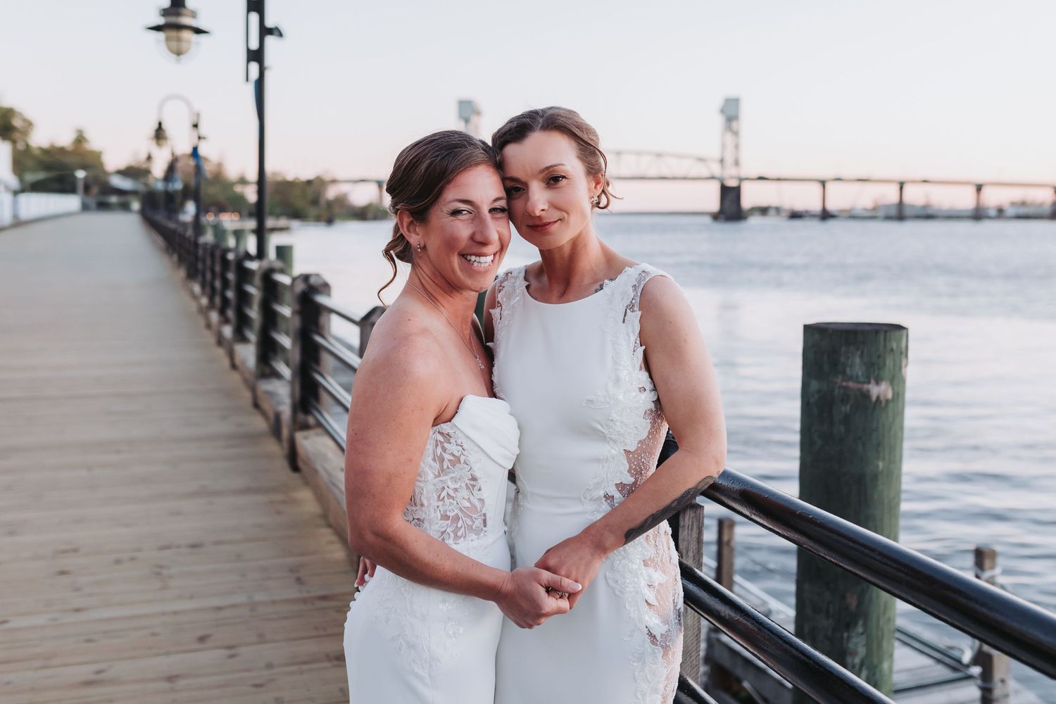 Two smiling brides in white wedding dresses pose together on a riverside boardwalk at golden hour.