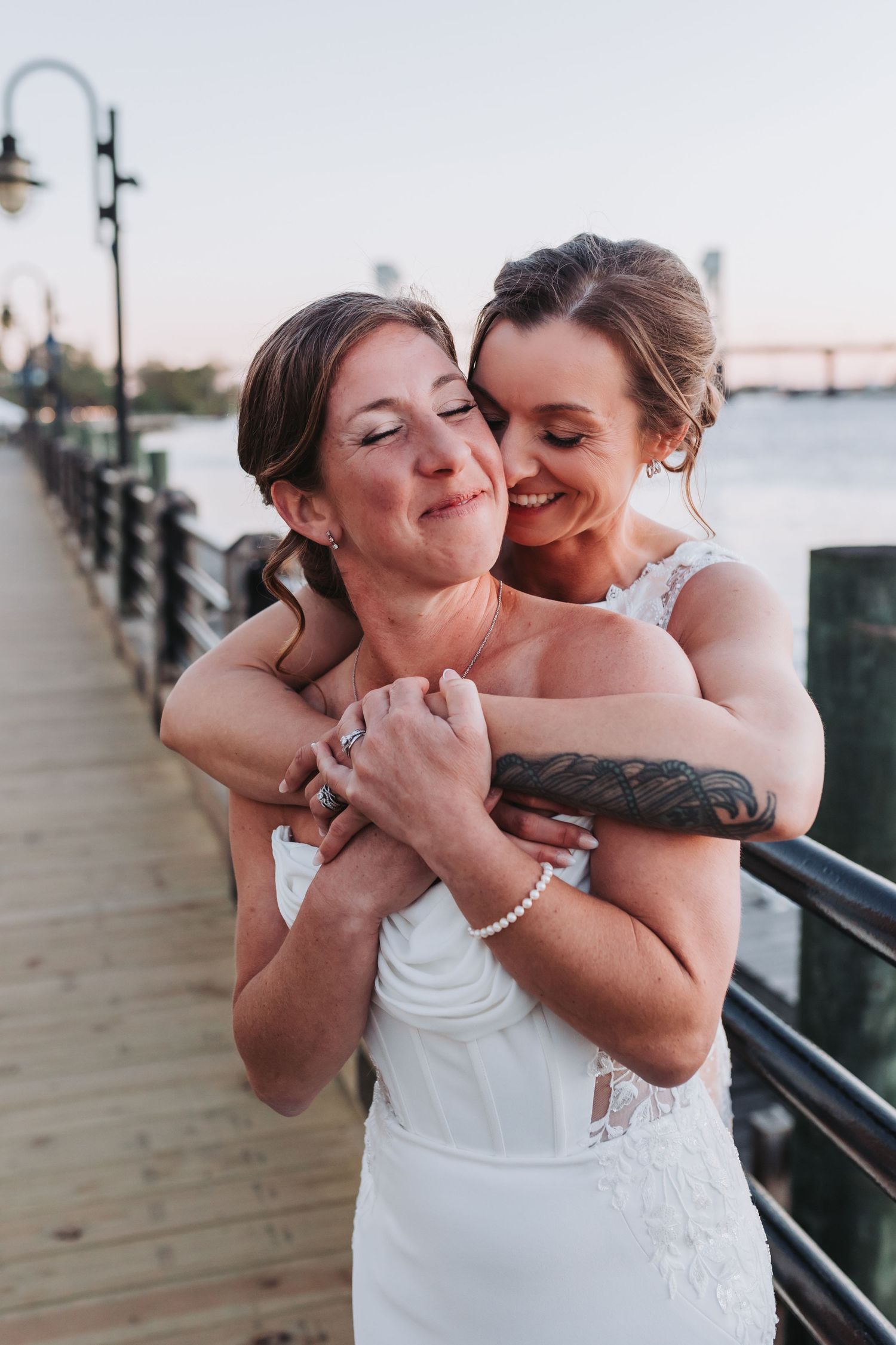 Two brides share a joyful embrace on a waterfront pier at golden hour sunset.