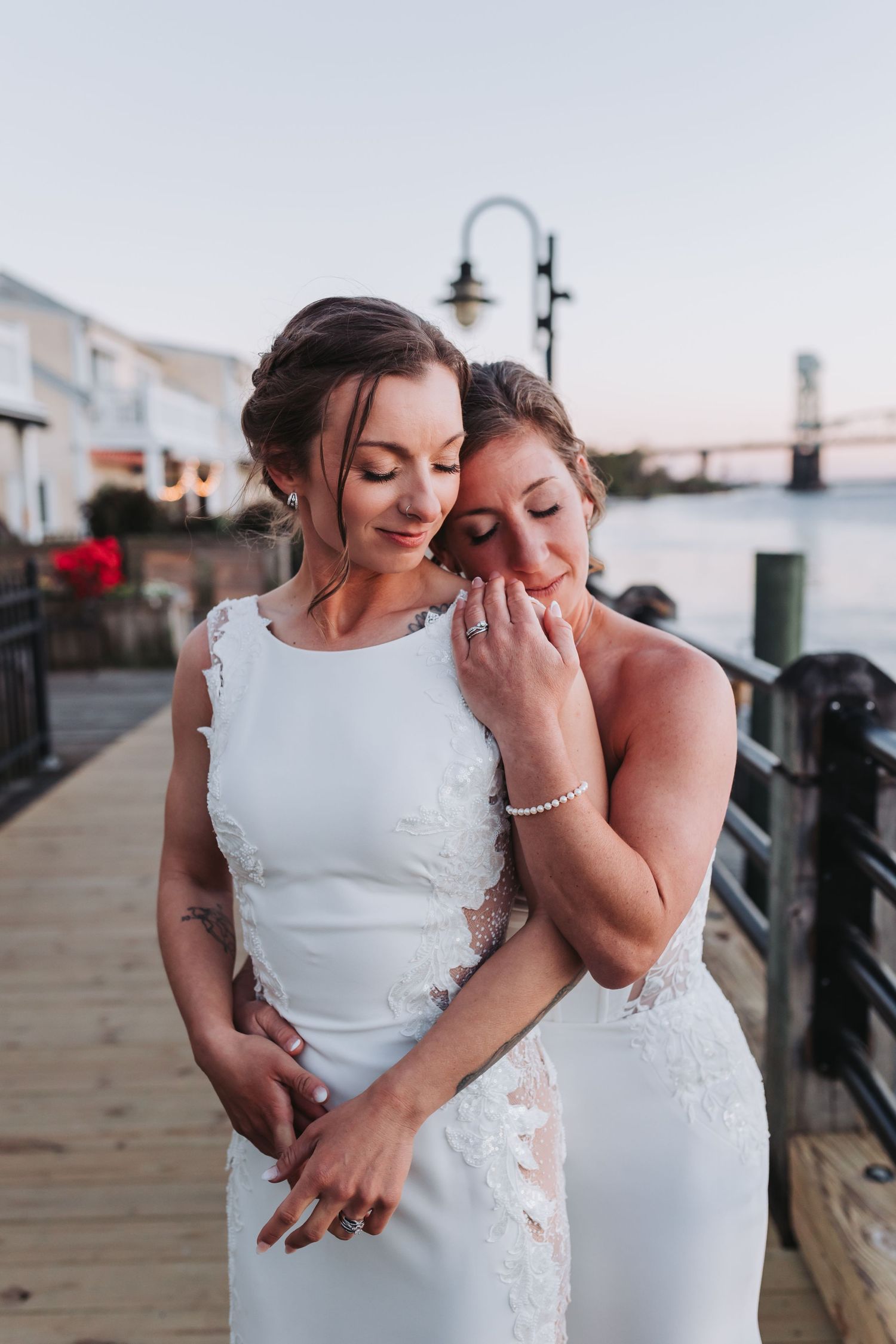 Two brides in white wedding dresses share an emotional embrace on a waterfront boardwalk at dusk.