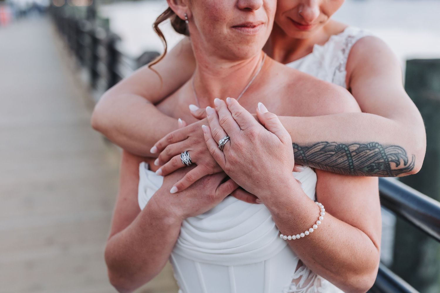 Two brides embracing, showing off wedding rings and pearl bracelet, one with a floral tattoo sleeve, in a romantic outdoor setting.