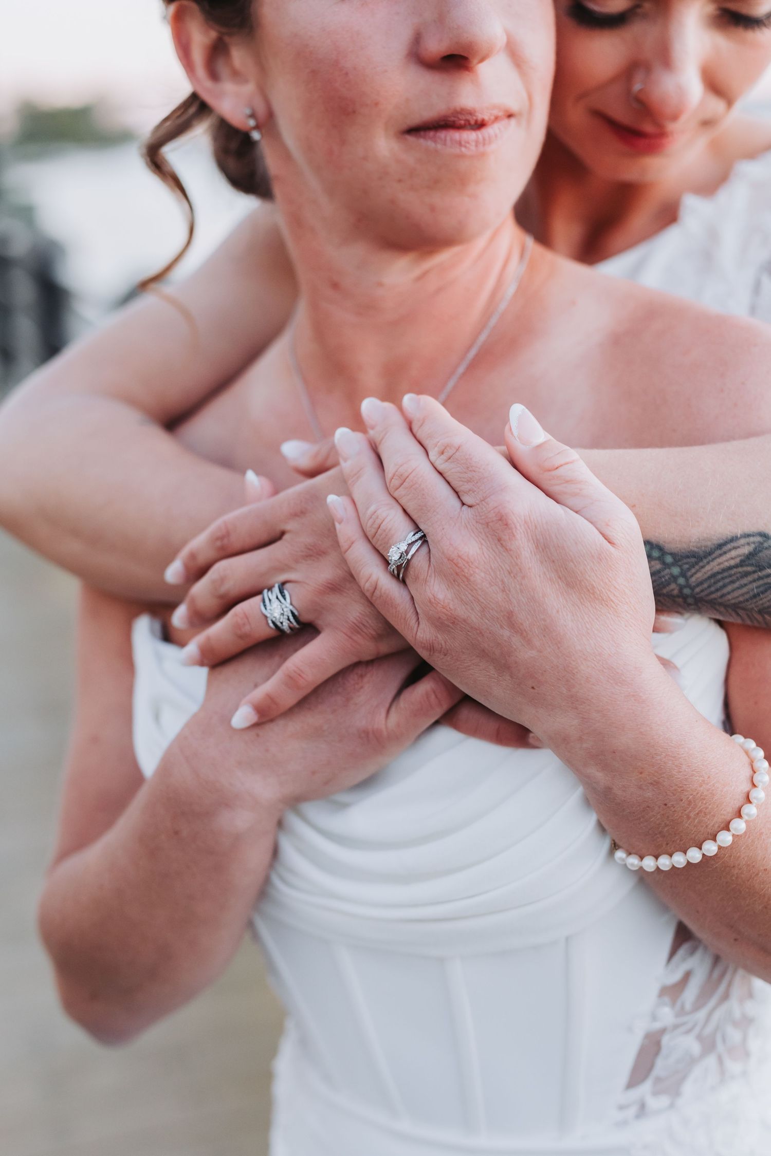 Two brides display matching wedding rings, hands clasped together, one wearing a white gown and pearl bracelet.