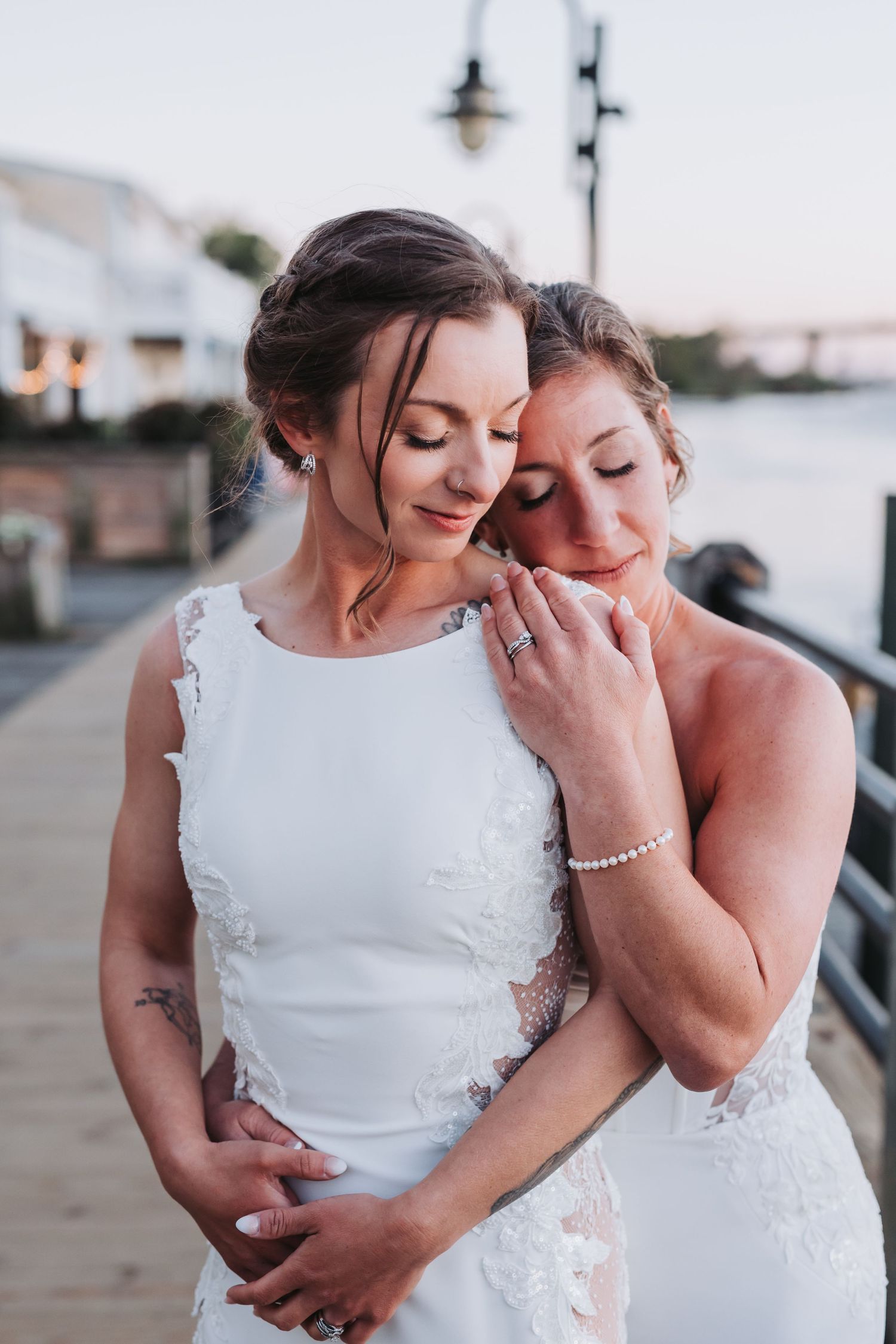 Two brides in white lace wedding gowns share an intimate embrace on a waterfront boardwalk at sunset.