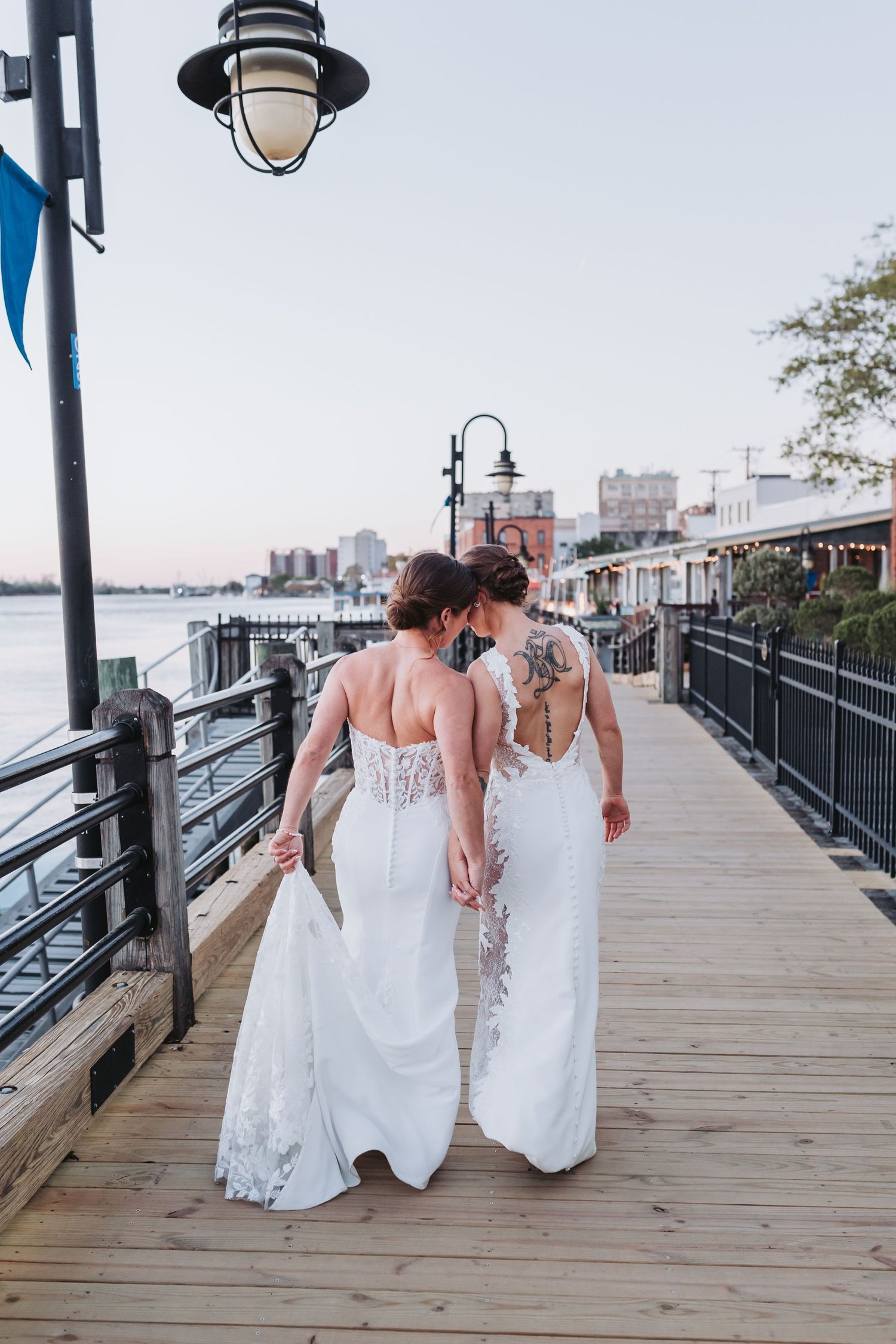 Two brides in elegant white wedding gowns walk hand in hand along a scenic waterfront boardwalk.