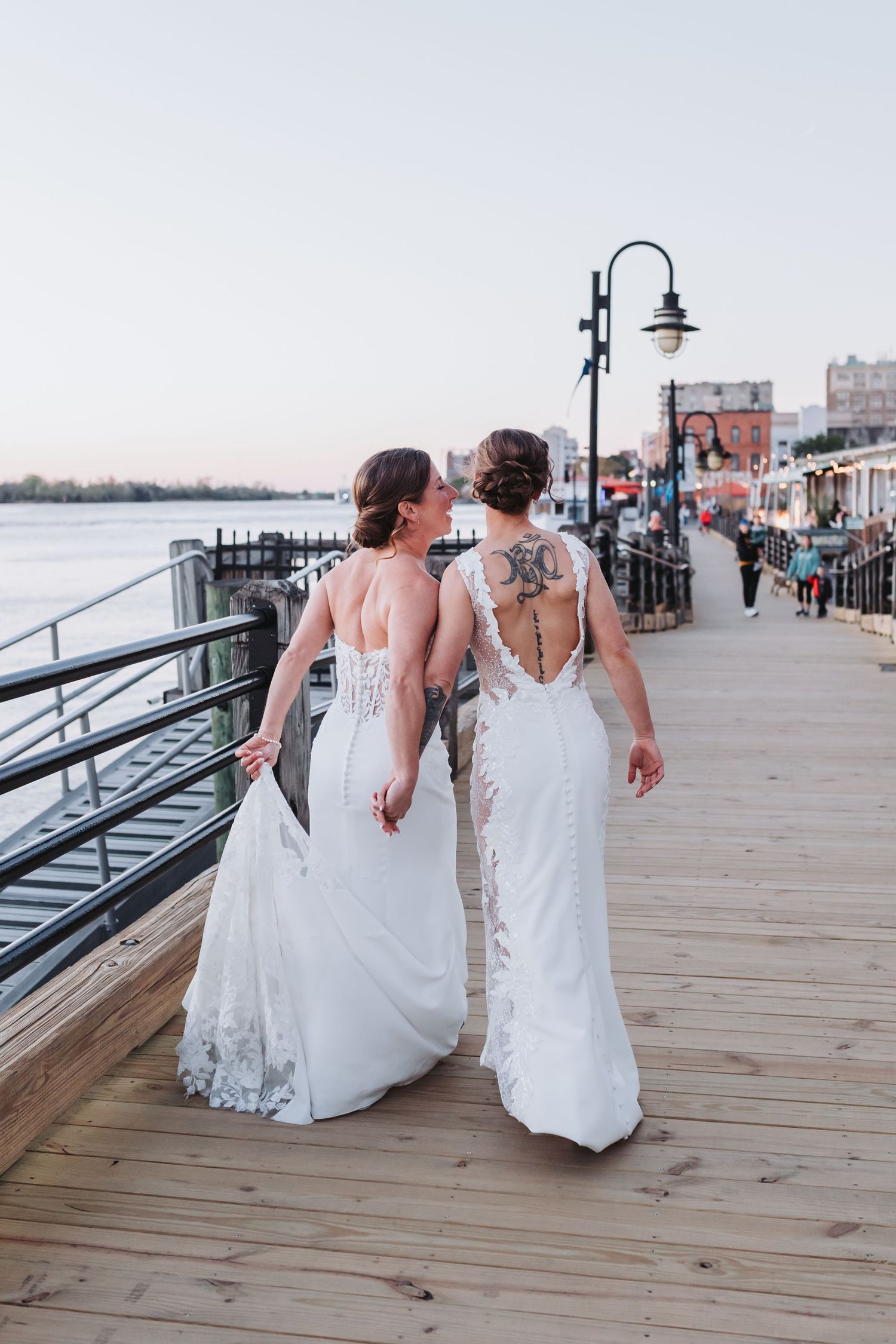 Two brides in white gowns with tattoos walk along a waterfront boardwalk on their wedding day.