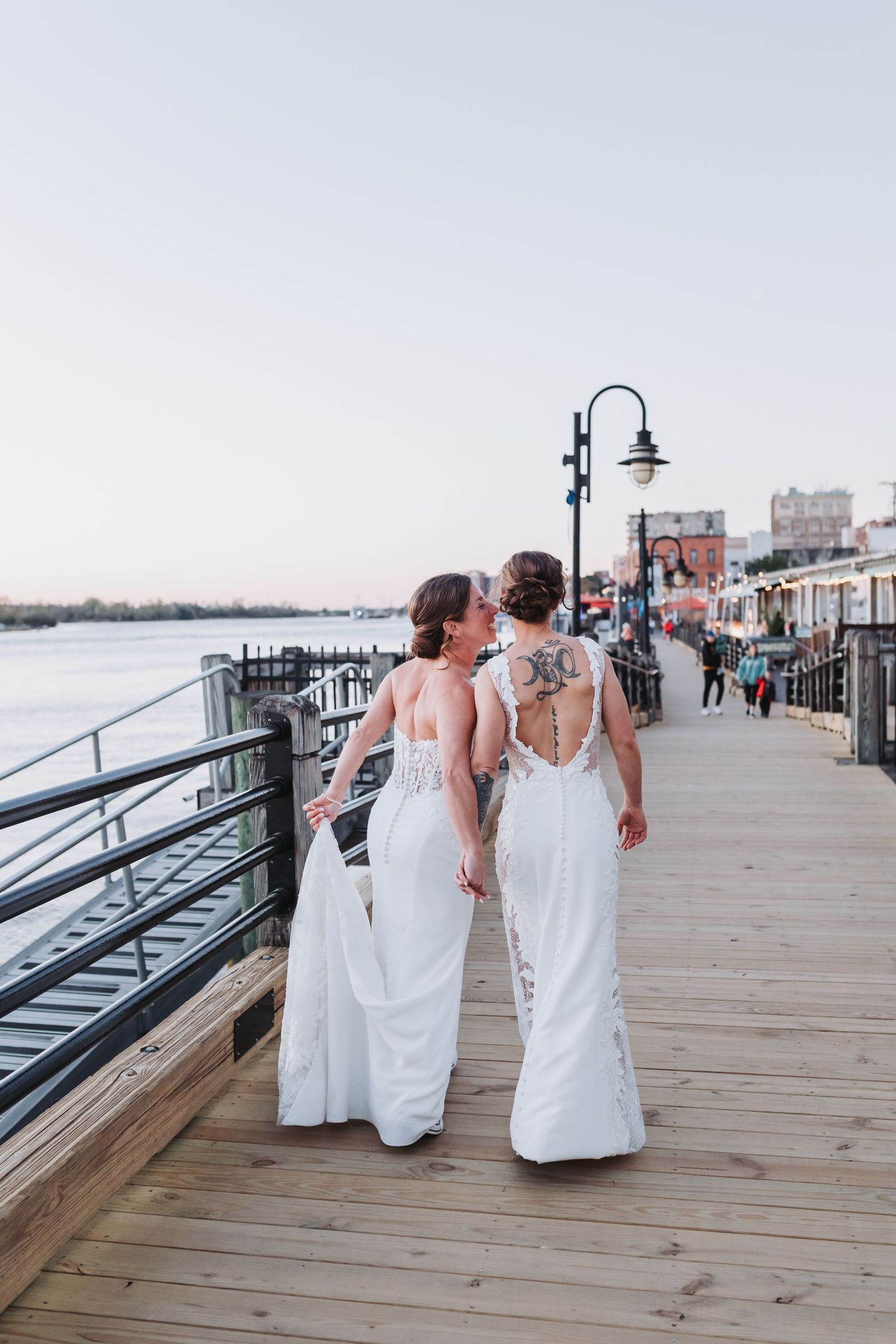 Two brides in white lace gowns walk hand-in-hand along a waterfront boardwalk at sunset.