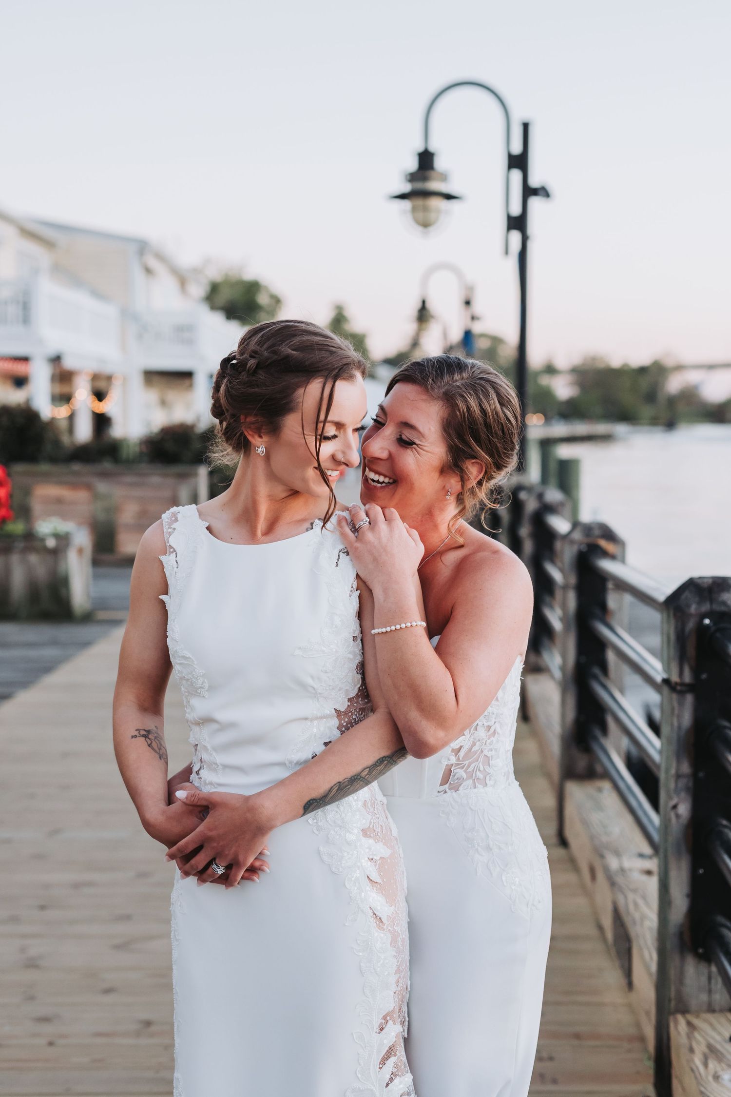 Two brides in white wedding gowns share a joyful moment on a waterfront dock at sunset.