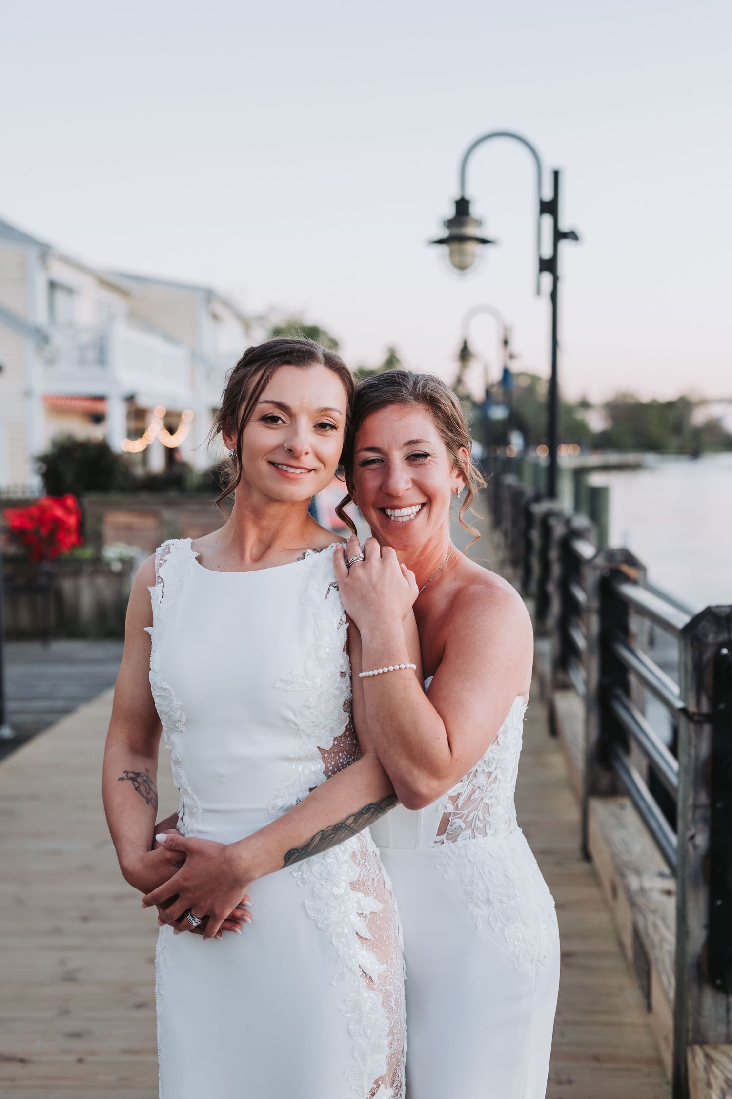 Two brides in white lace wedding gowns embrace on a waterfront boardwalk at dusk, smiling joyfully.