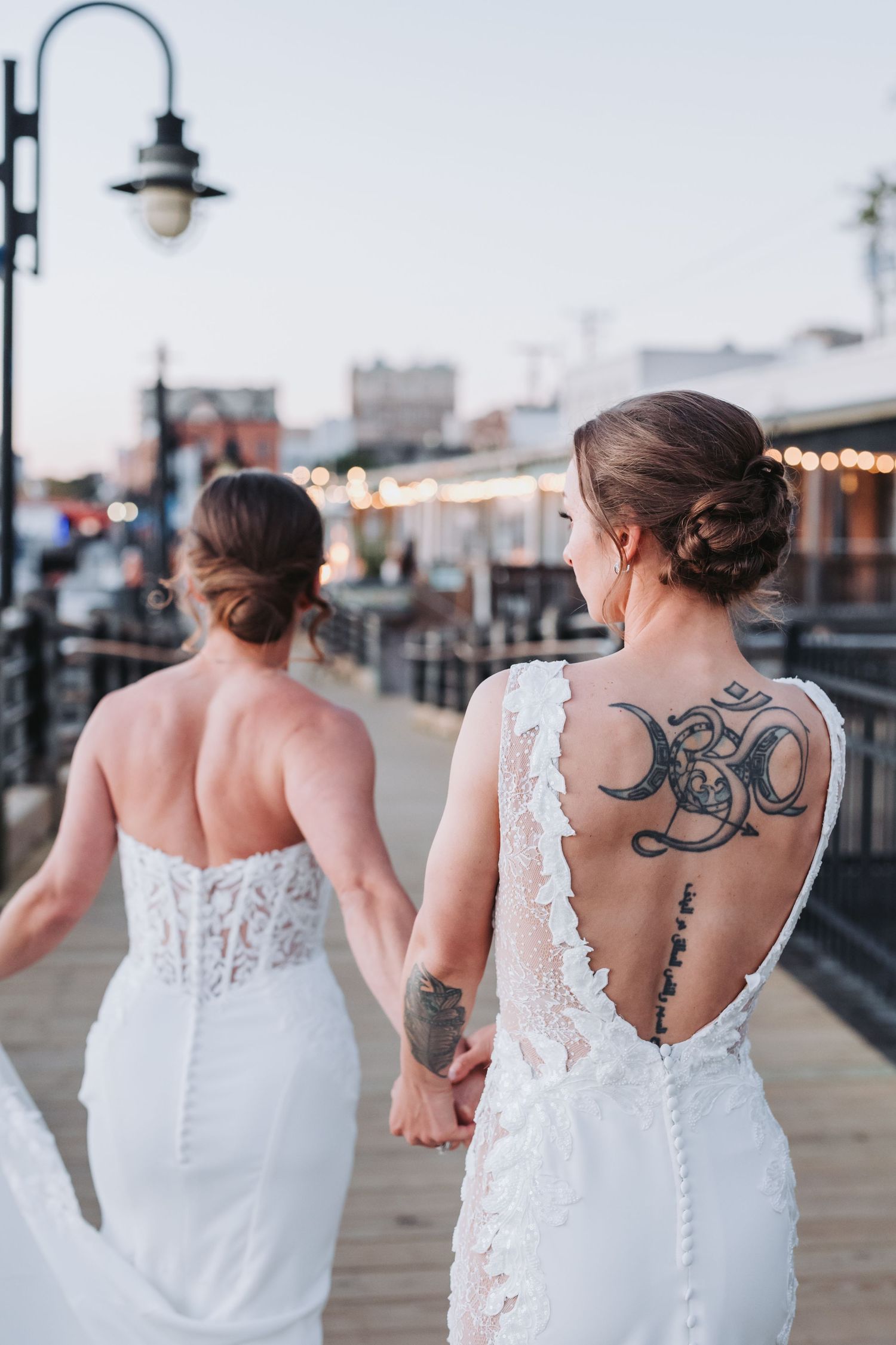 Two brides walk hand in hand on a boardwalk, one featuring elegant back tattoos visible through her lace gown.