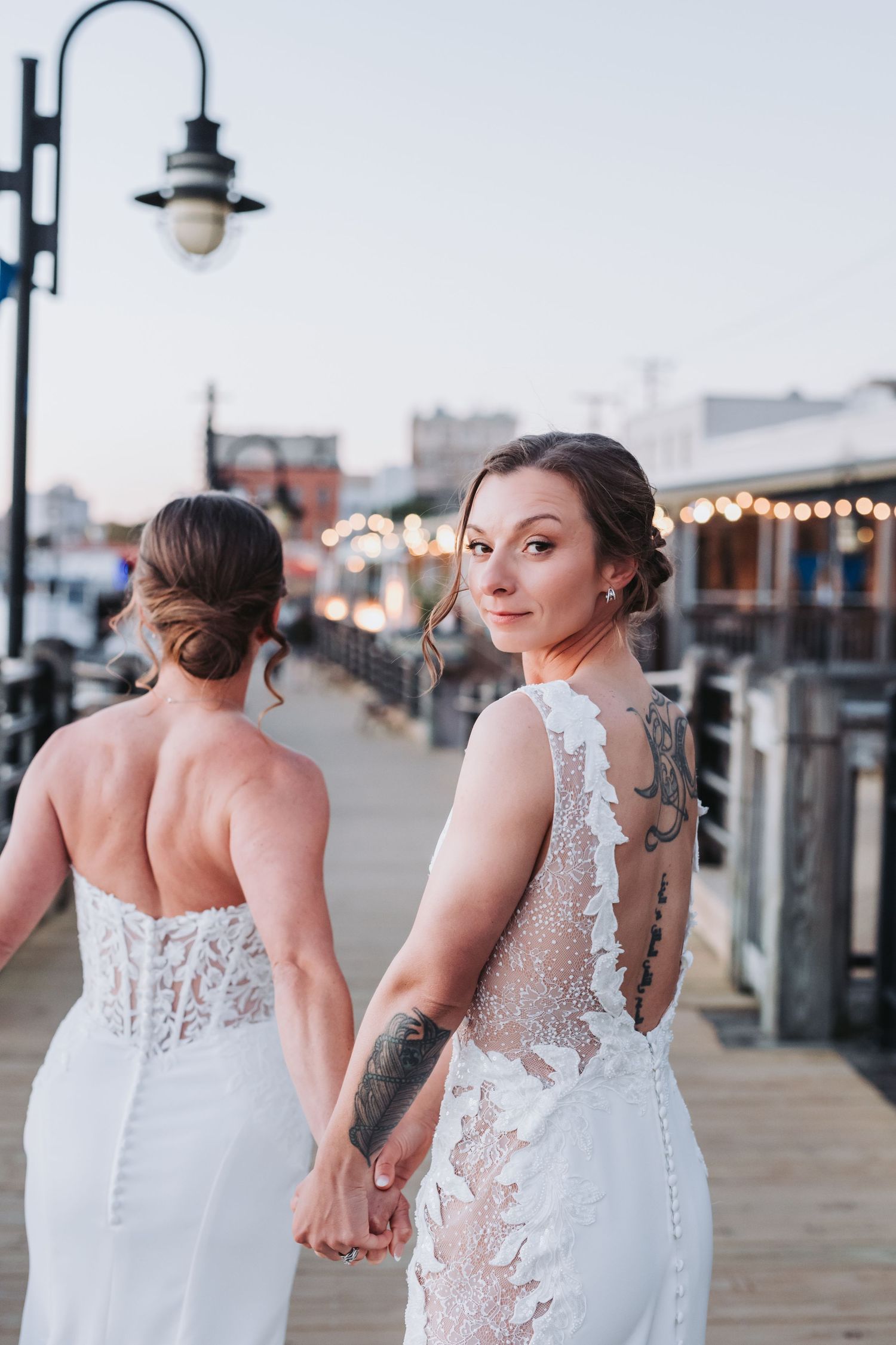 Two brides in lace wedding gowns hold hands on a waterfront boardwalk at dusk, one glancing back at the camera.