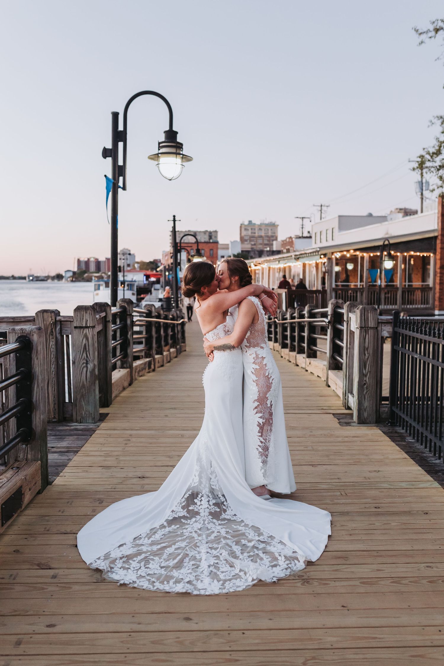 Two brides in white lace wedding gowns embrace on a wooden boardwalk pier at dusk near the waterfront.