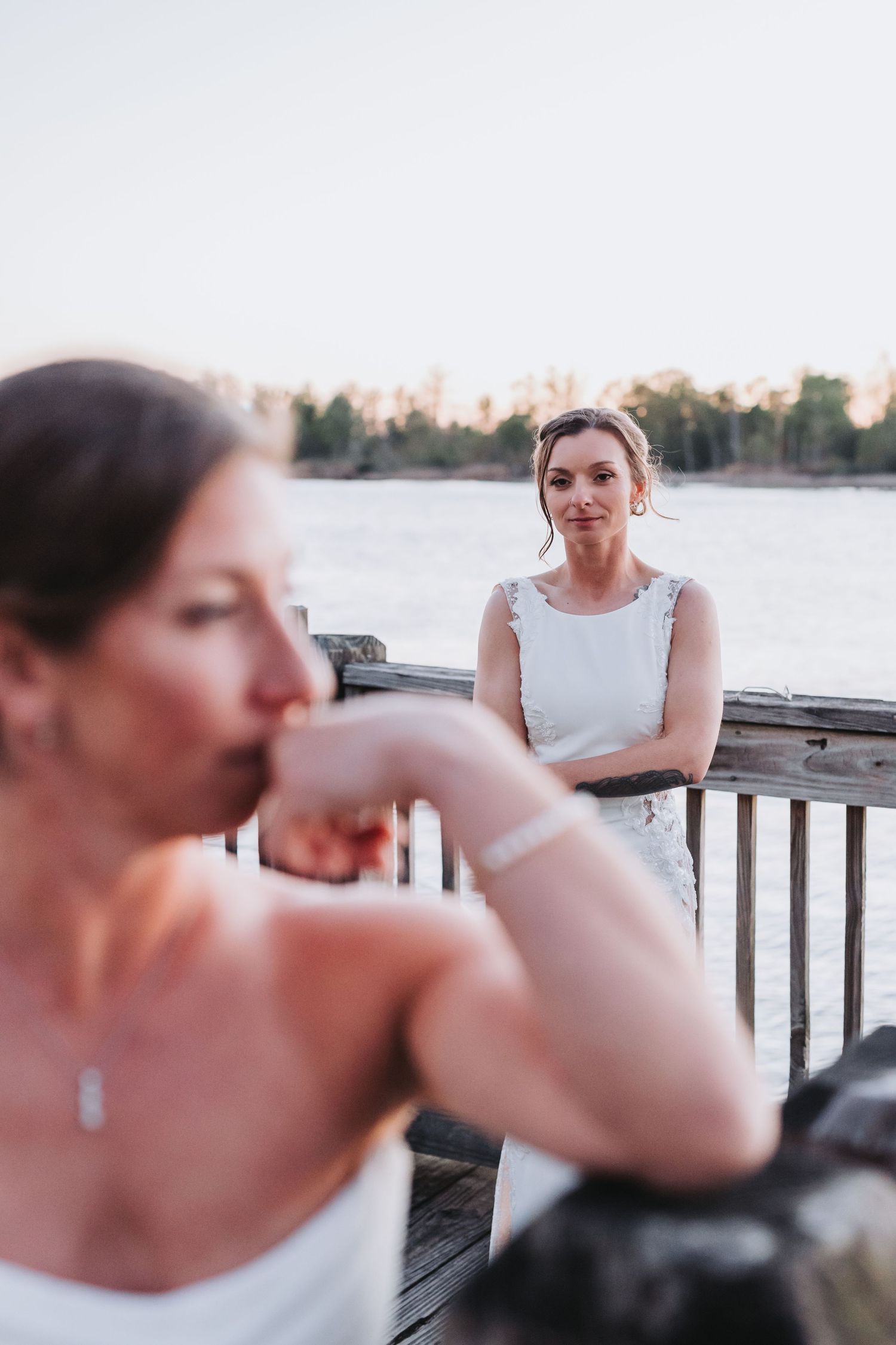 Two brides in white wedding dresses share a romantic moment on a waterfront dock at sunset.