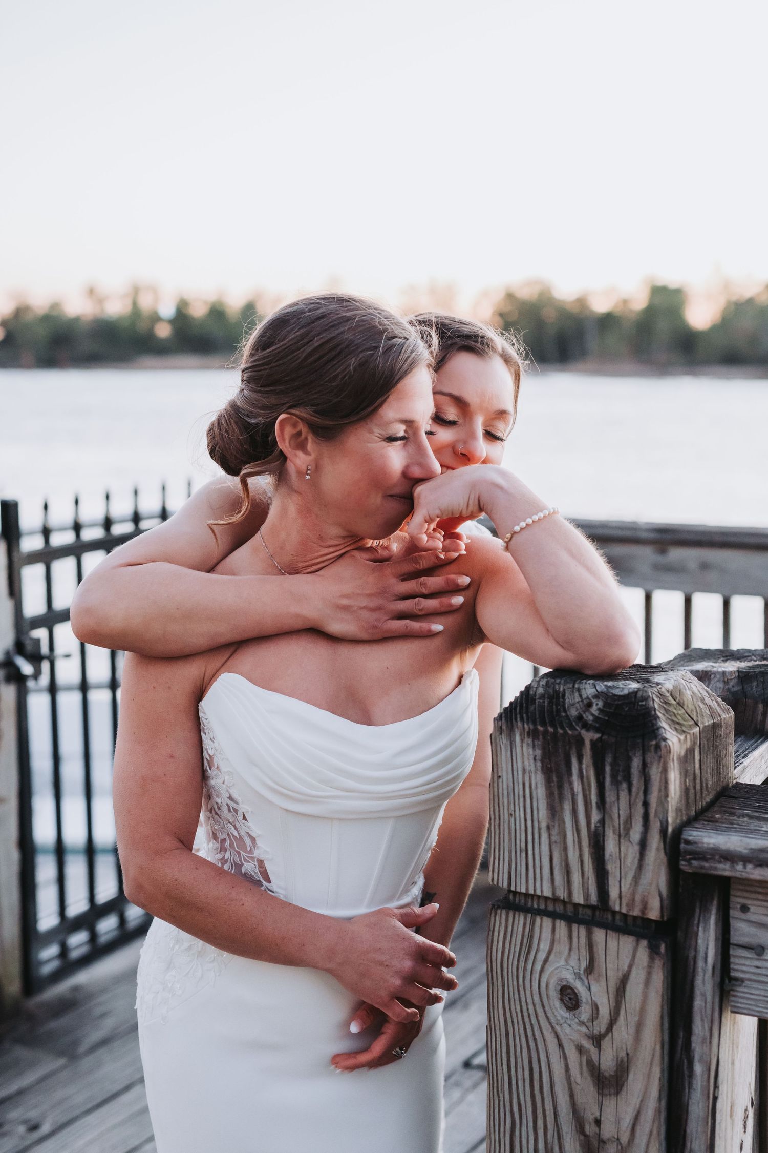 Two brides share a tender embrace on a wooden dock overlooking a serene waterway at dusk.