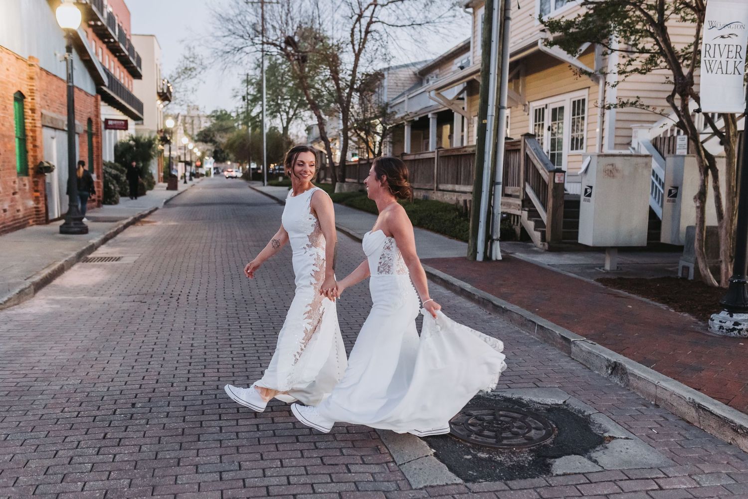 Two brides in white wedding gowns walk hand-in-hand down a charming brick street lined with colorful buildings.