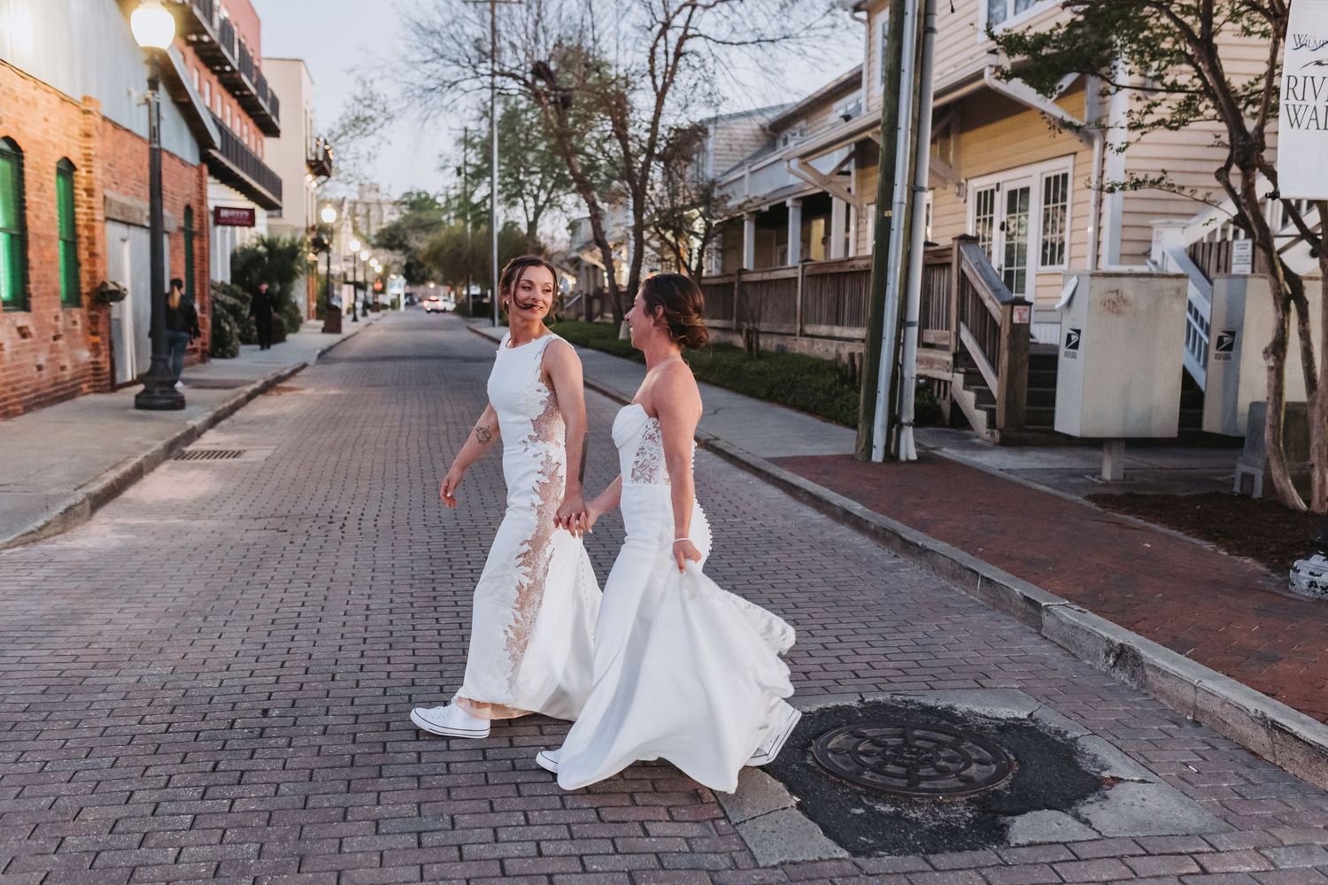 Two brides in white wedding gowns walk hand-in-hand on a brick street in a charming historic town.