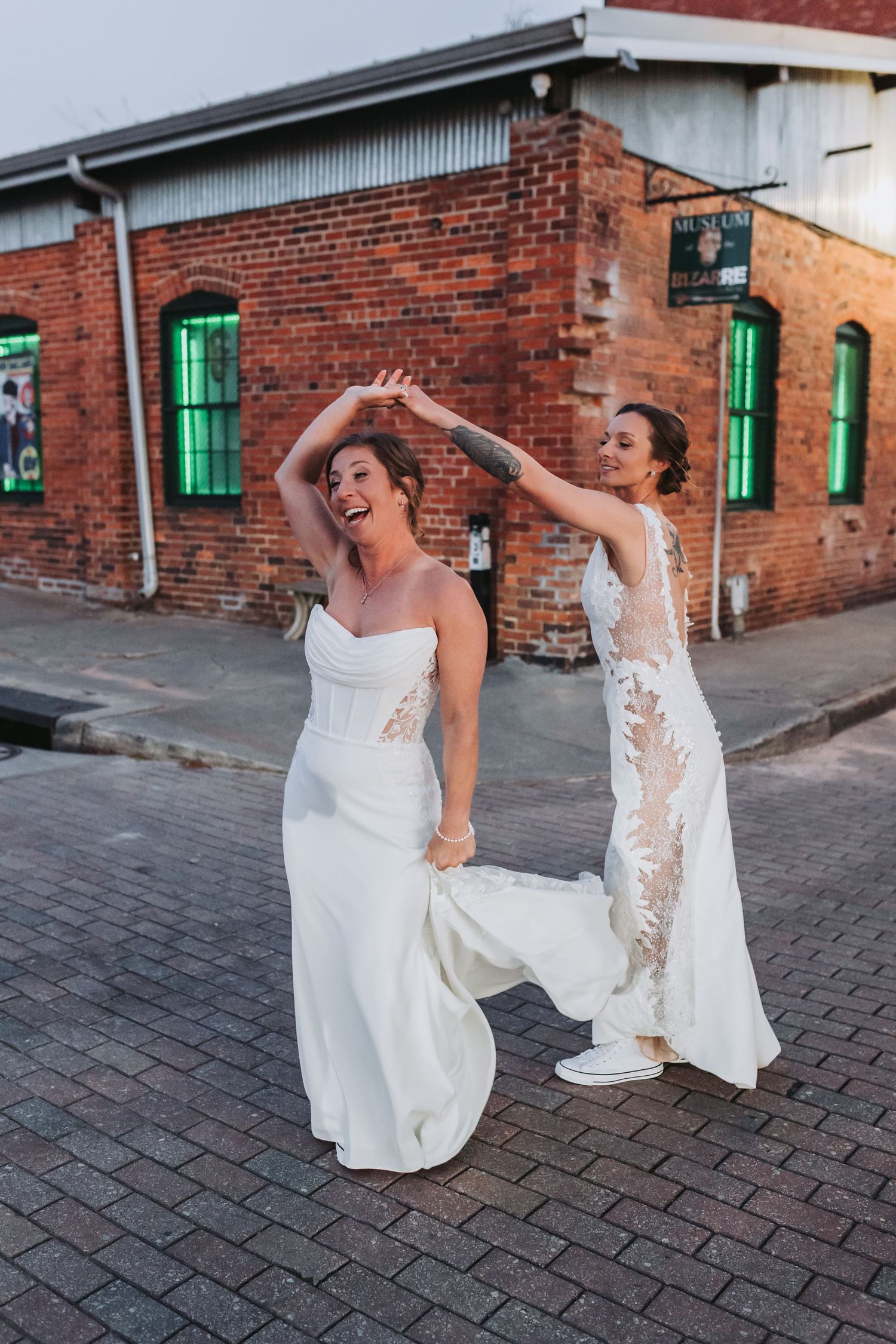 Two brides in elegant white gowns dance joyfully on a cobblestone street at golden hour.
