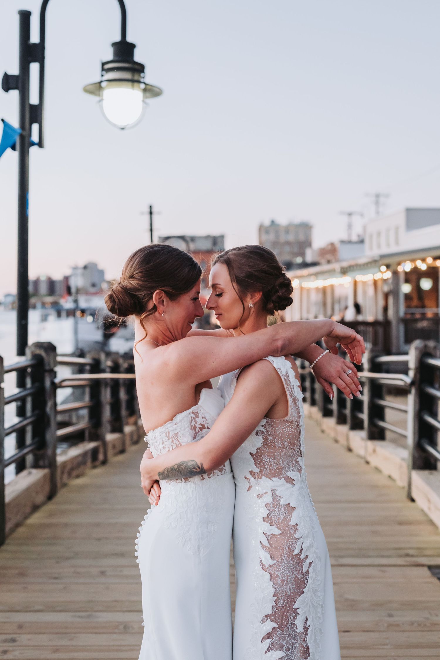 Two brides in lace wedding gowns share an intimate embrace on a waterfront pier at dusk.