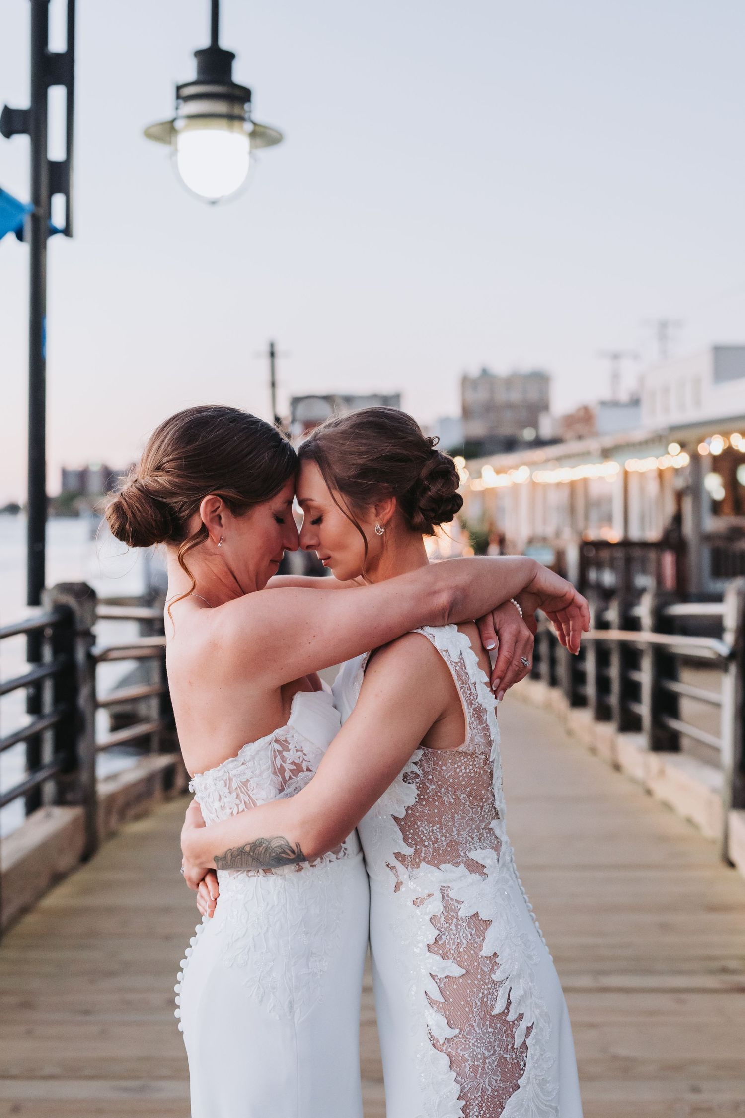 Two brides in lace wedding gowns embrace tenderly on a waterfront pier at sunset.