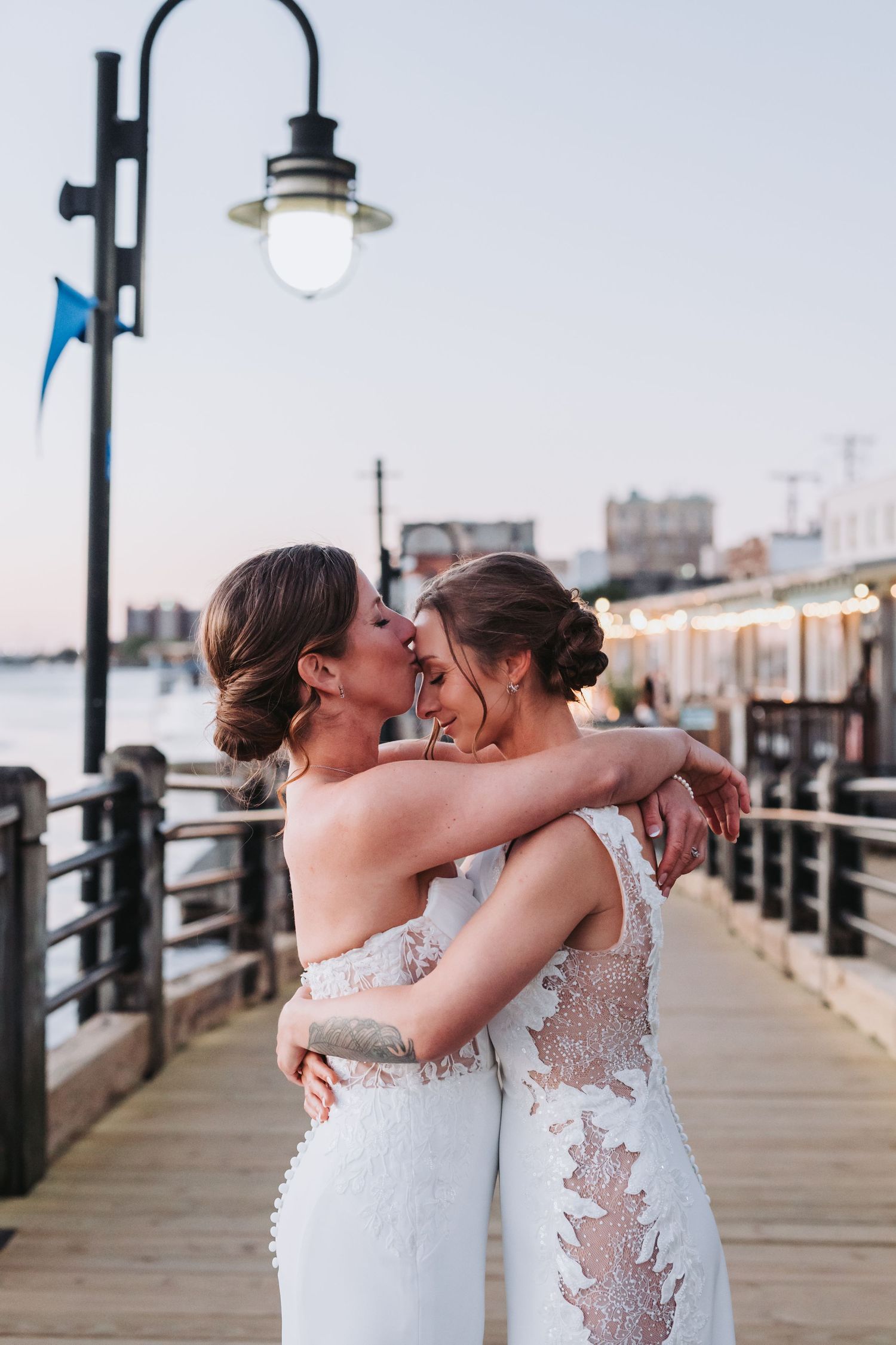 Two brides in lace wedding gowns embrace and kiss on a waterfront pier at dusk.