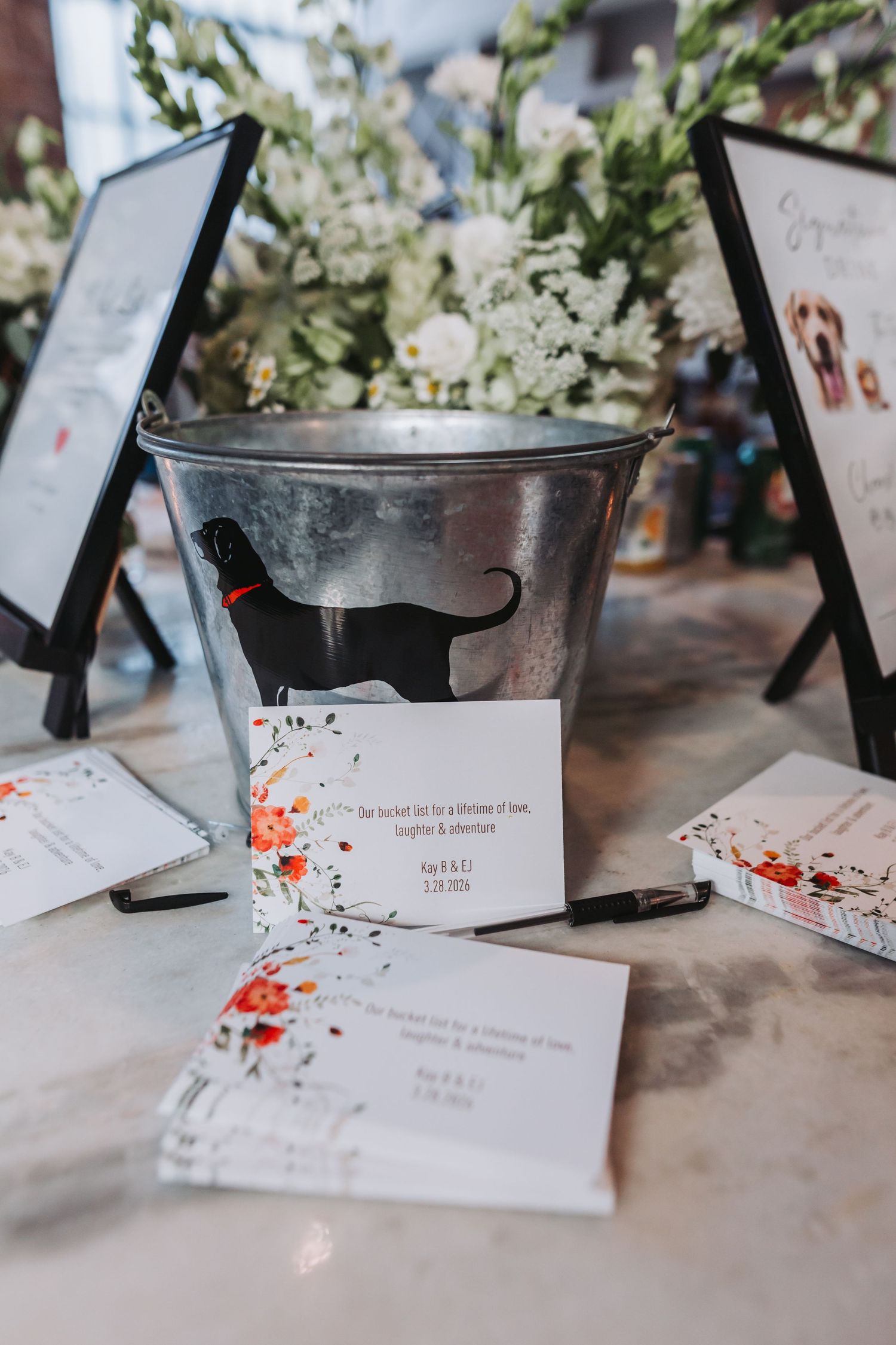 Silver bucket with black cat silhouette surrounded by wedding bucket list cards and floral arrangements at a reception.