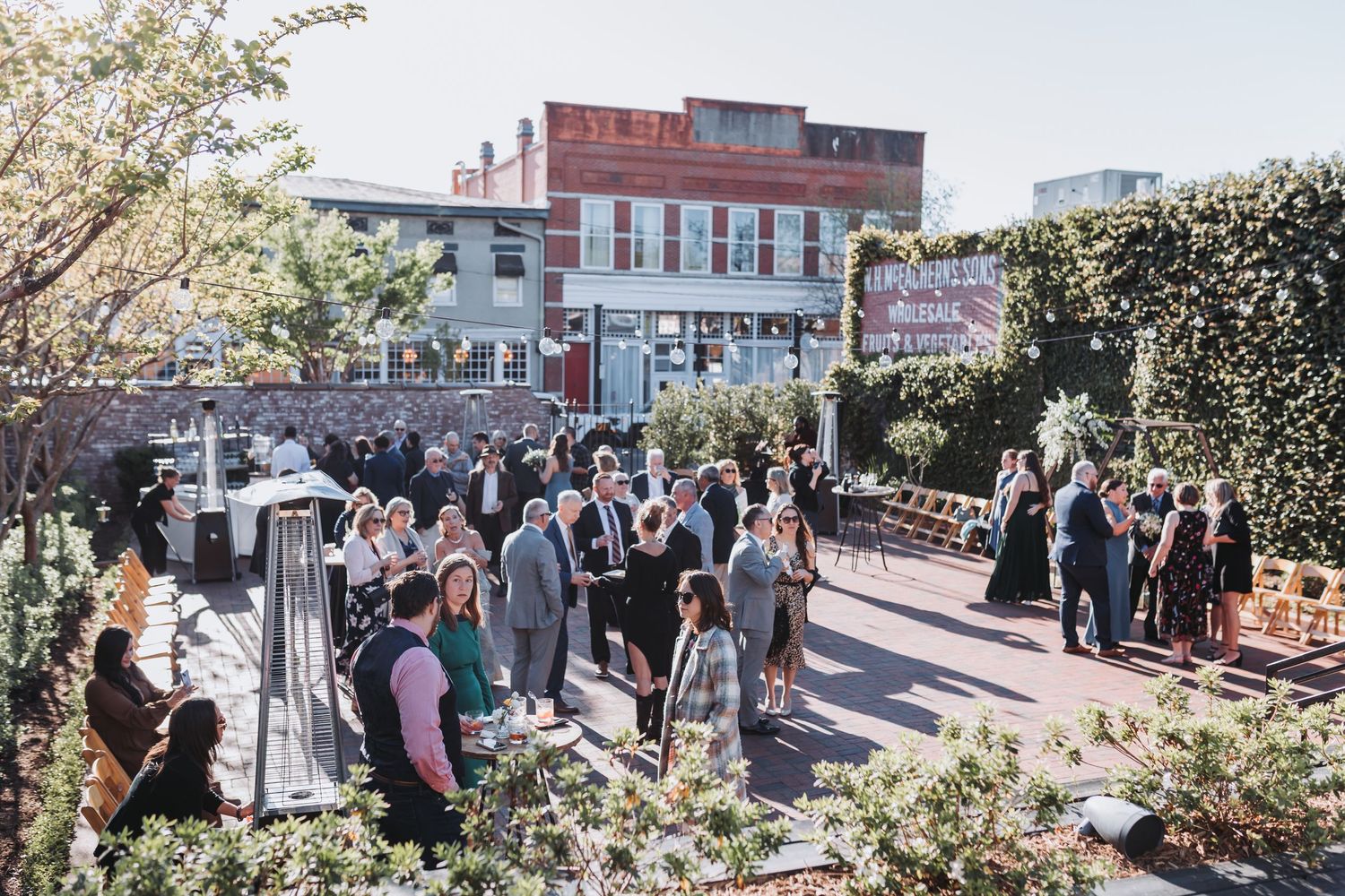 Outdoor wedding cocktail hour with guests mingling on a sunny urban patio surrounded by greenery and brick buildings.