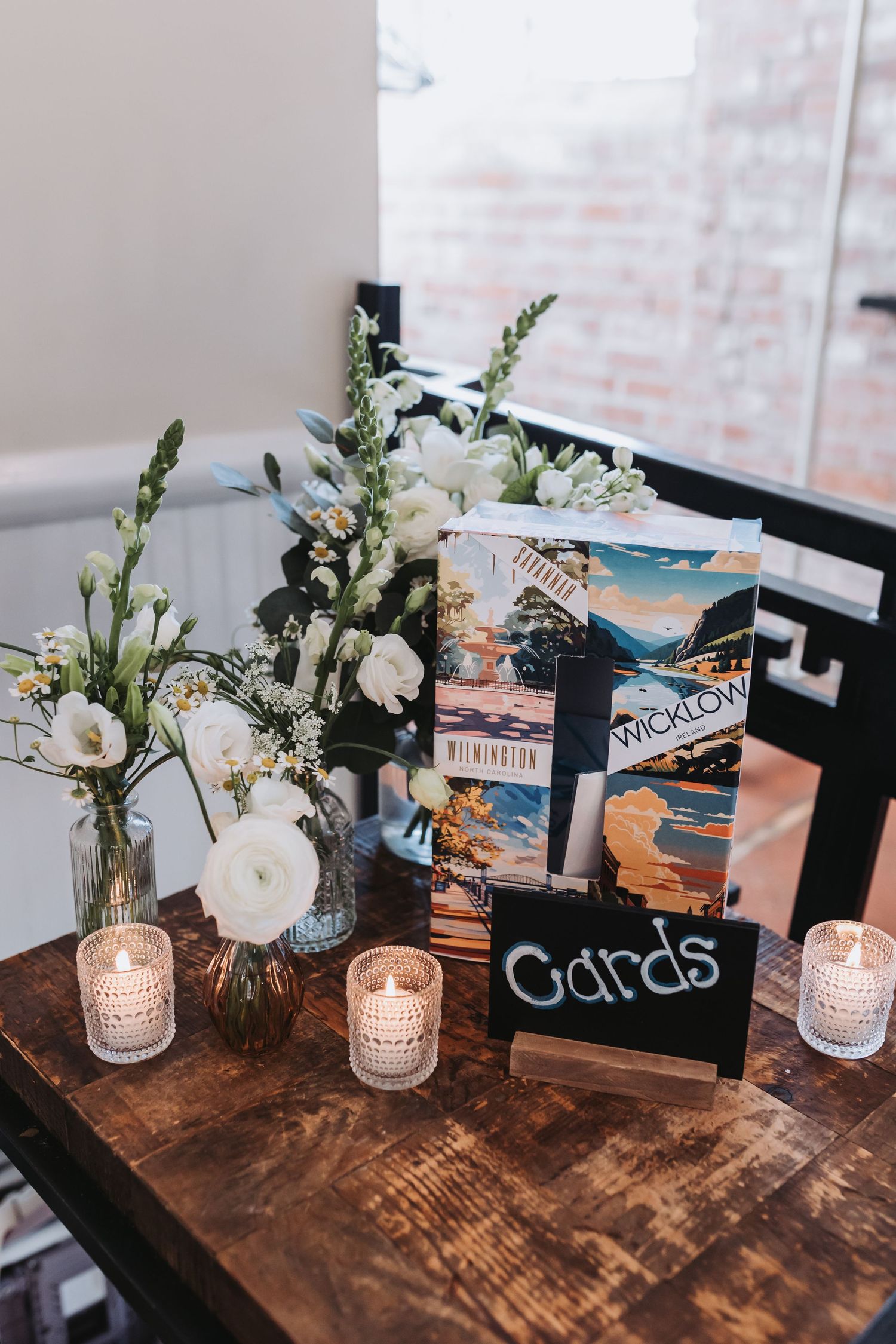 Wedding card table with white floral arrangement, vintage travel postcards, and glowing votive candles on rustic wood.