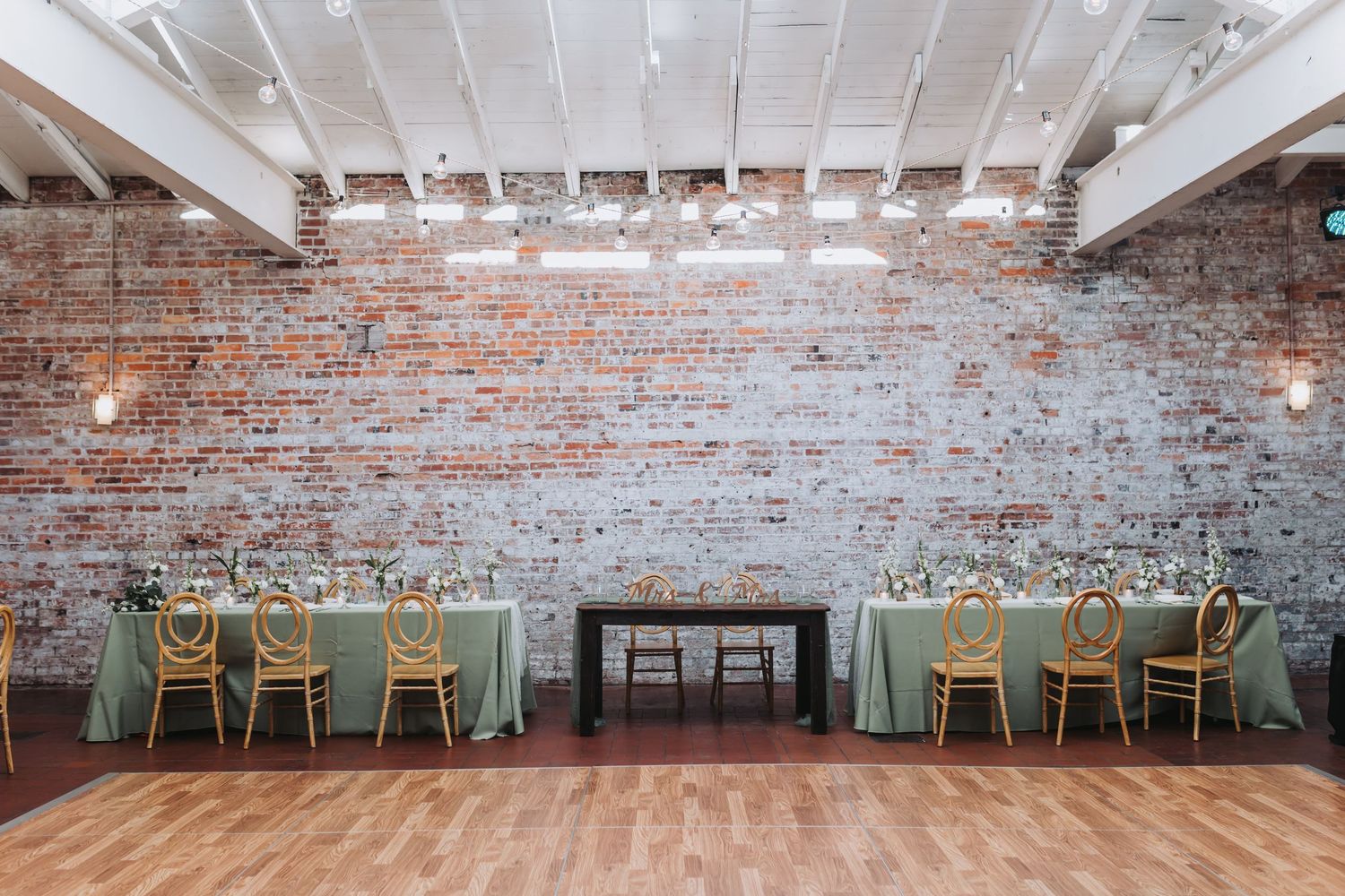 Elegant wedding reception venue with exposed brick wall, sage green tablecloths, and warm string lights overhead.