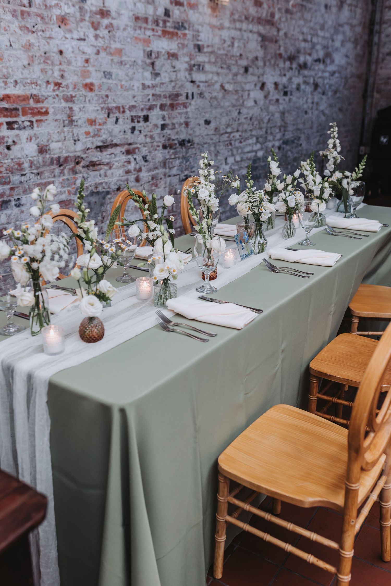 Elegant wedding head table with sage green linen, white floral arrangements, and candles against an exposed brick wall.