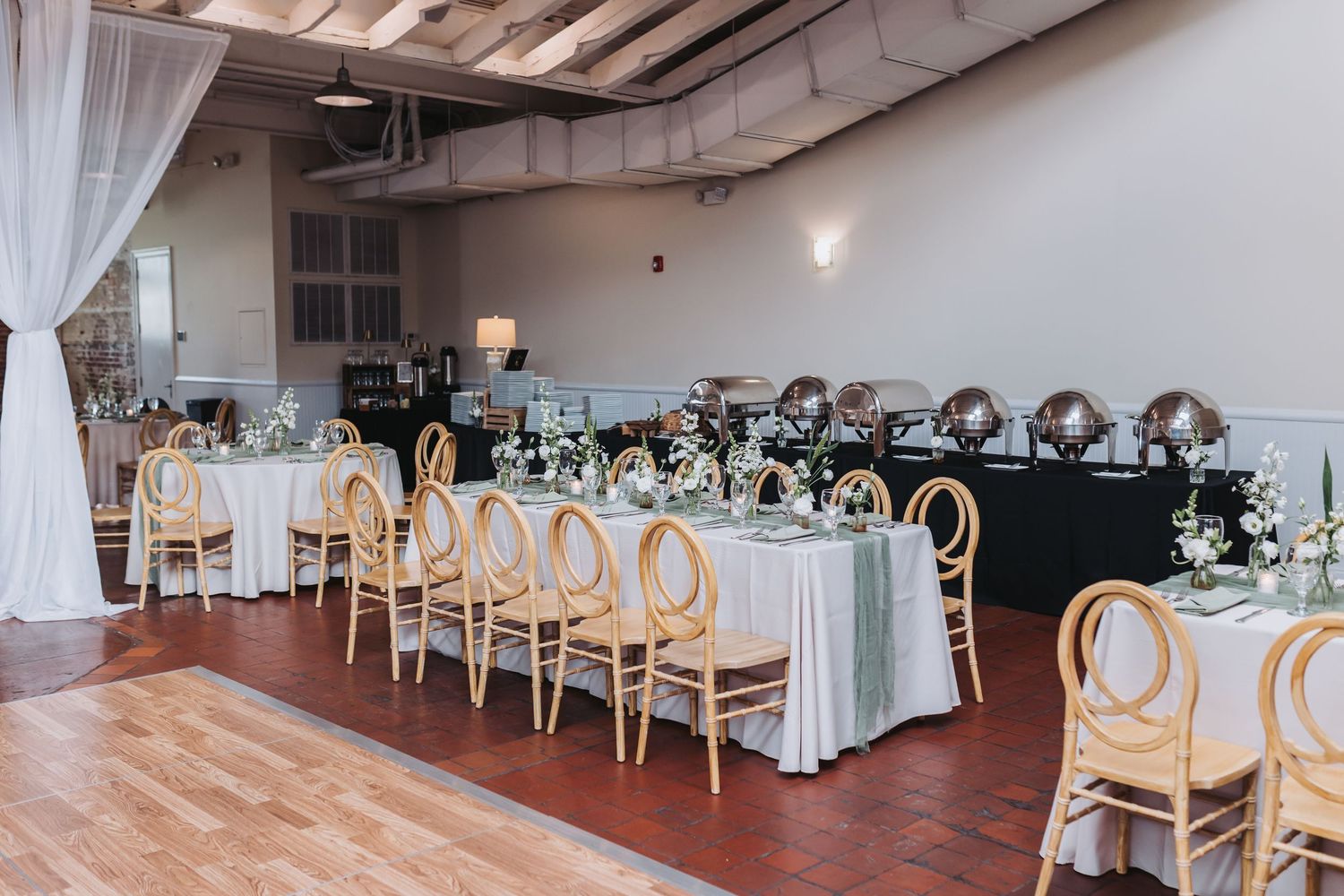 Elegant wedding reception hall with rattan chairs, white linen tables, floral centerpieces, and draped curtains on hardwood floors.