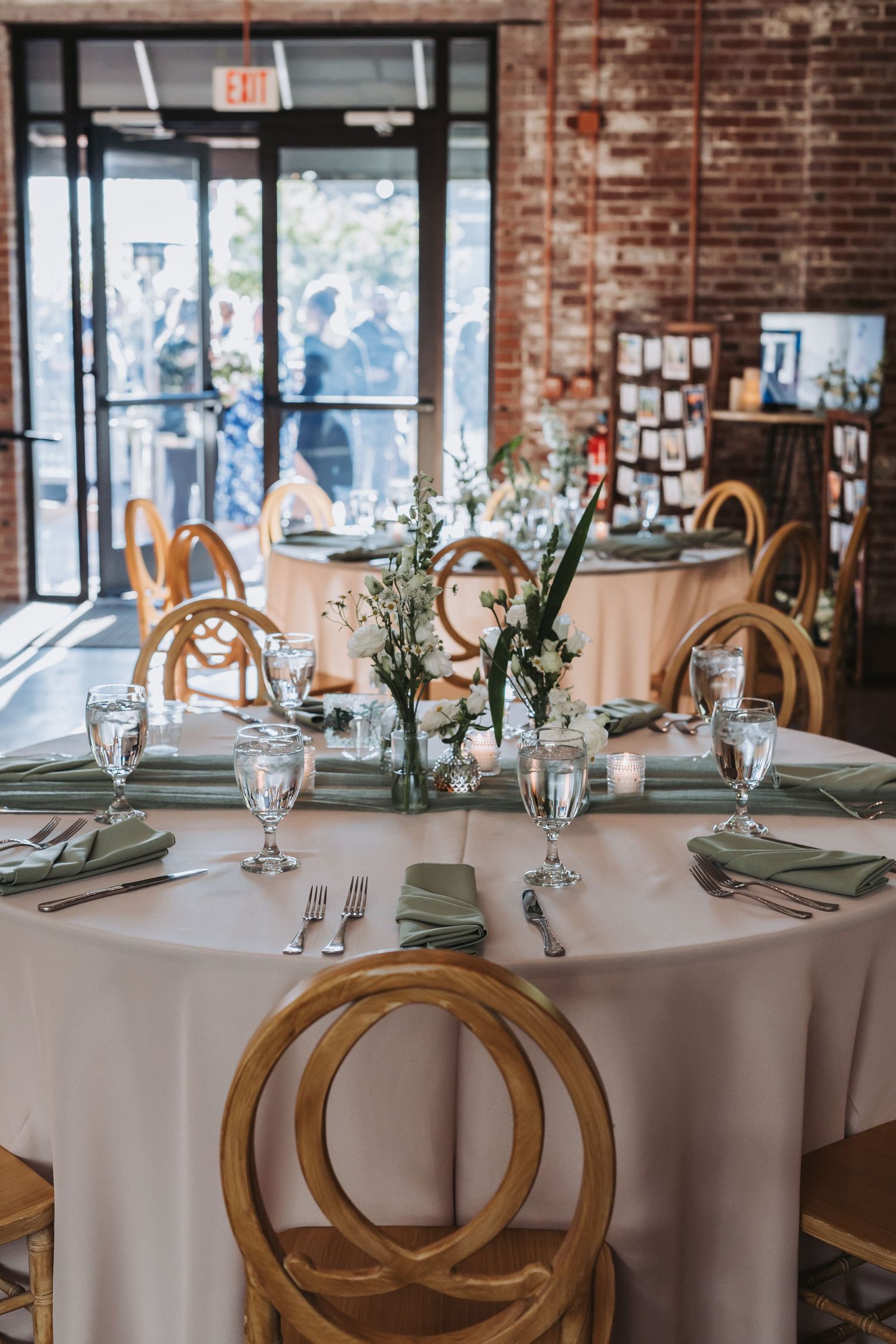 Round wedding reception tables with white linens, sage napkins, wood cross-back chairs, and delicate floral centerpieces.