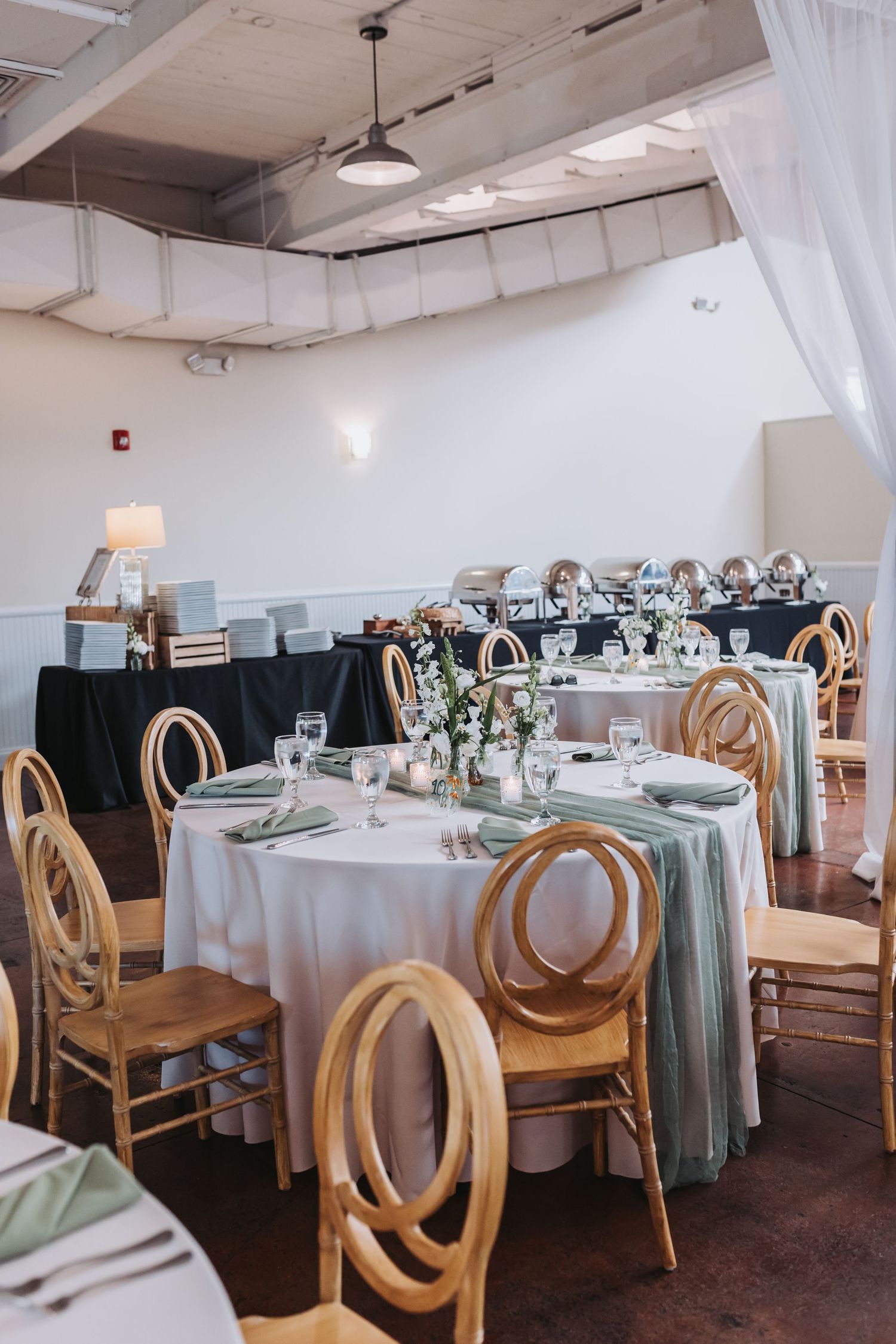 Elegant wedding reception with round tables, gold chairs, sage green napkins, and white floral centerpieces in bright venue.