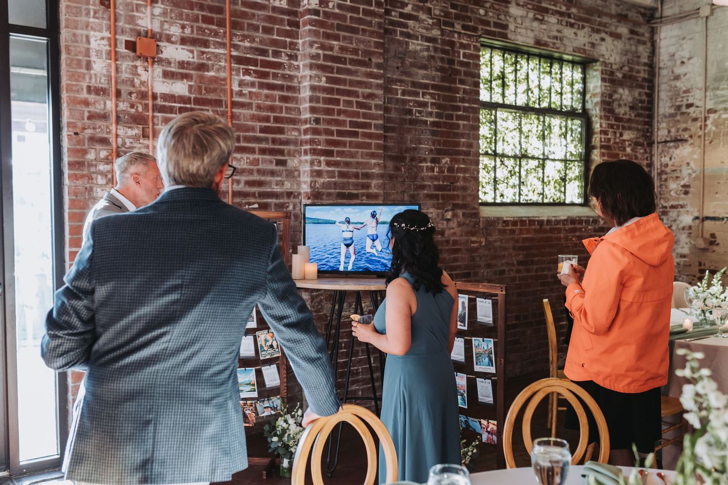 Wedding guests viewing a photo display on a TV monitor at a rustic brick wall reception venue.