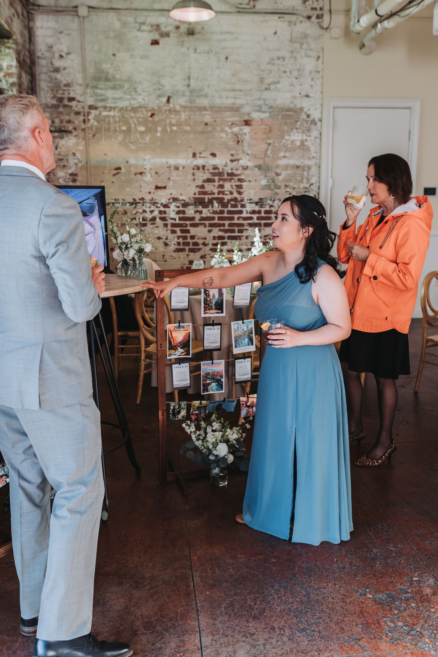 A bridesmaid in a blue gown reaches out to greet a guest in a gray suit at a rustic brick-walled wedding reception.