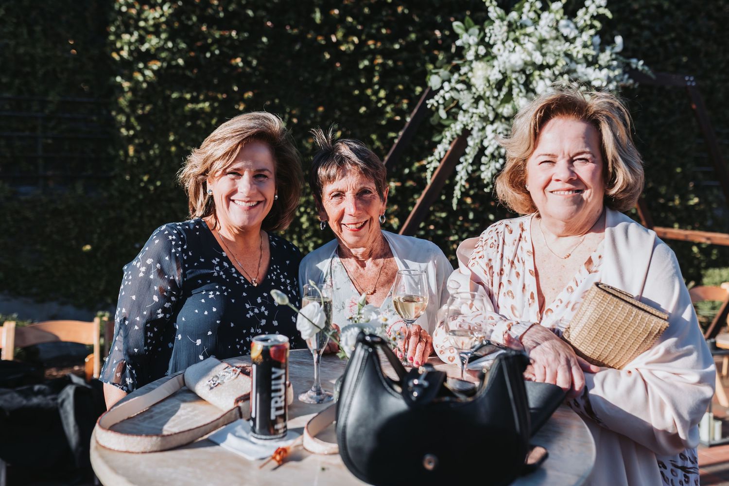 Three smiling women posing together at an outdoor wedding reception surrounded by white floral greenery.