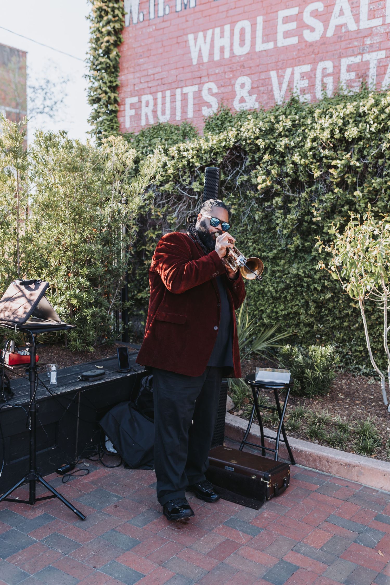 Musician in red velvet blazer playing trumpet outdoors near a wholesale fruits and vegetables brick building.