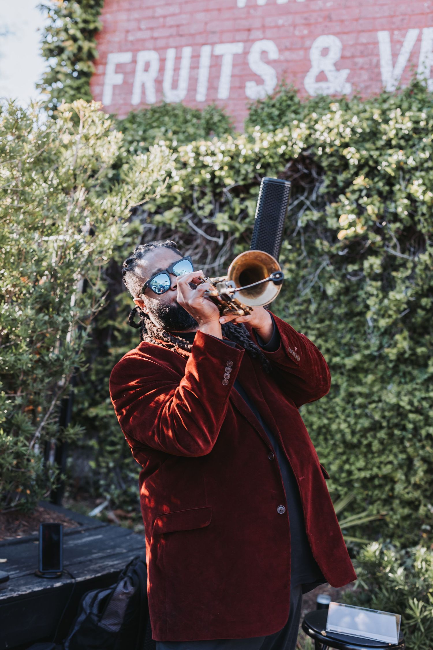 A musician in a red velvet blazer plays trumpet outdoors near a brick wall with a Fruits and Vegetables sign.