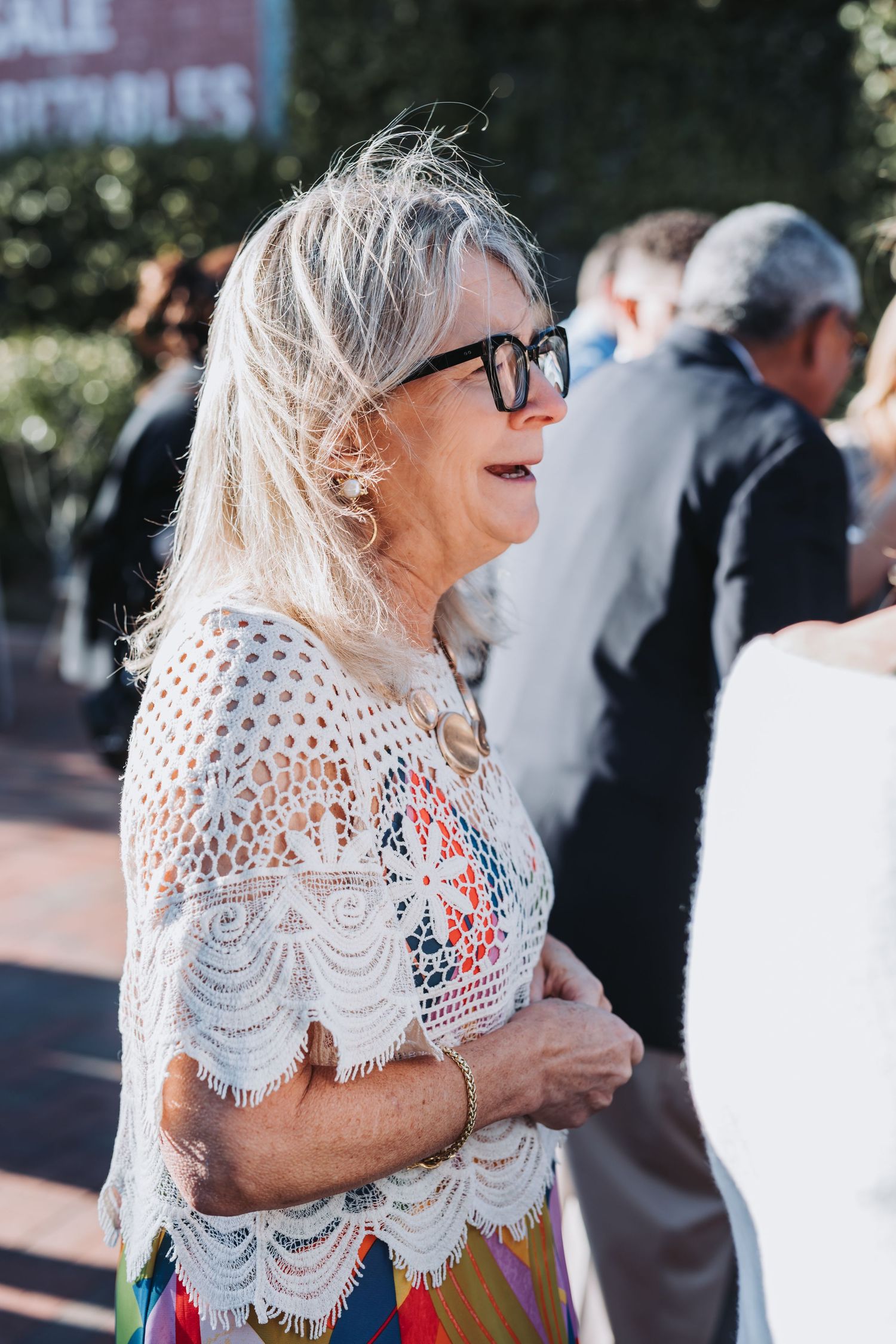 Older woman with blonde hair and black glasses wearing a white lace top at an outdoor wedding event.