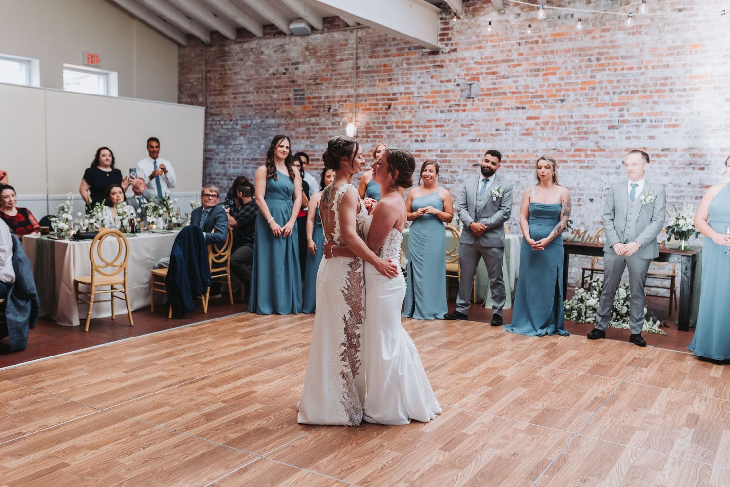 Two brides share their first dance surrounded by guests in a rustic brick wedding venue.