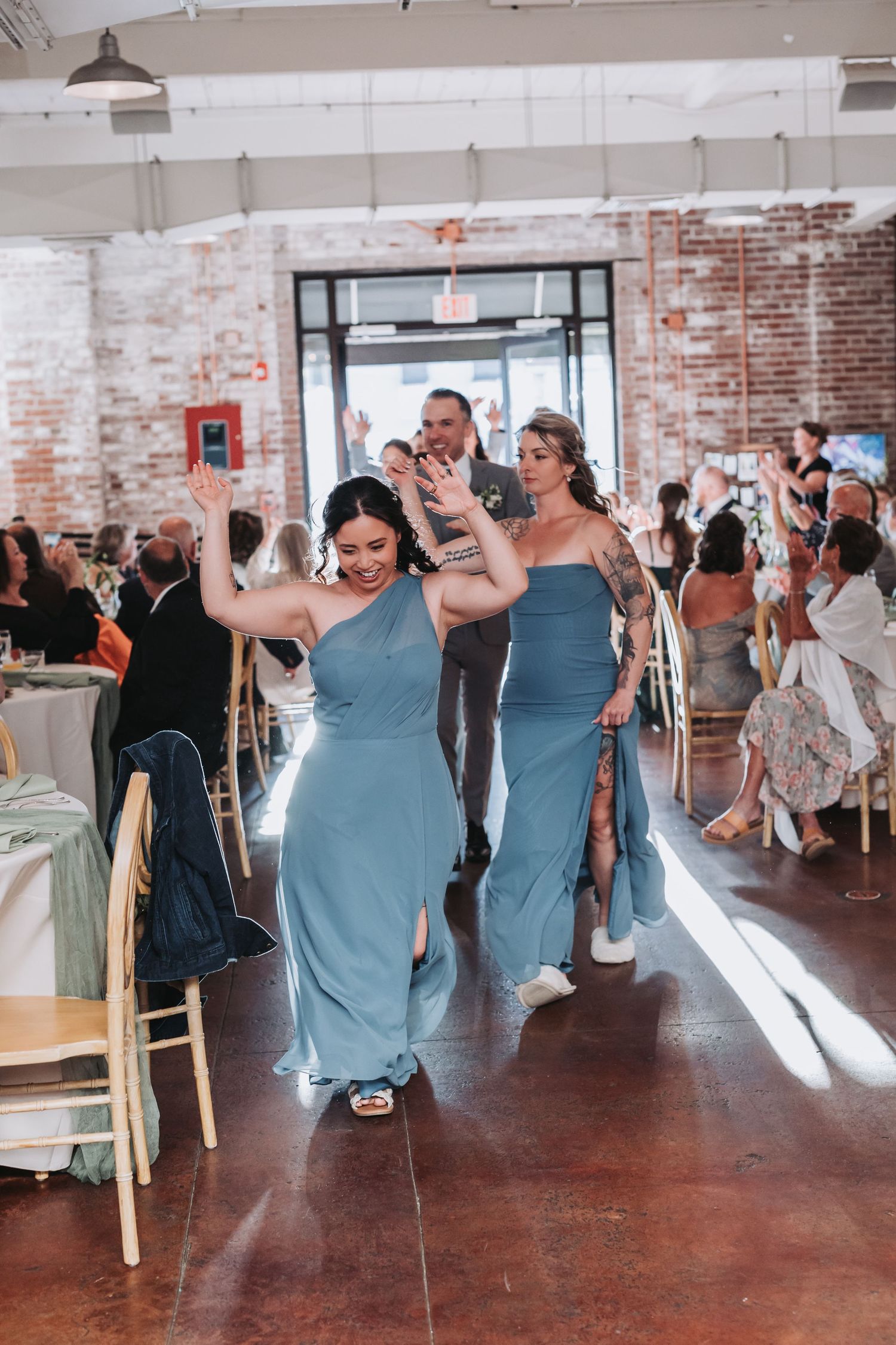 Bridesmaids in blue gowns make grand entrance at wedding reception in brick-walled venue.