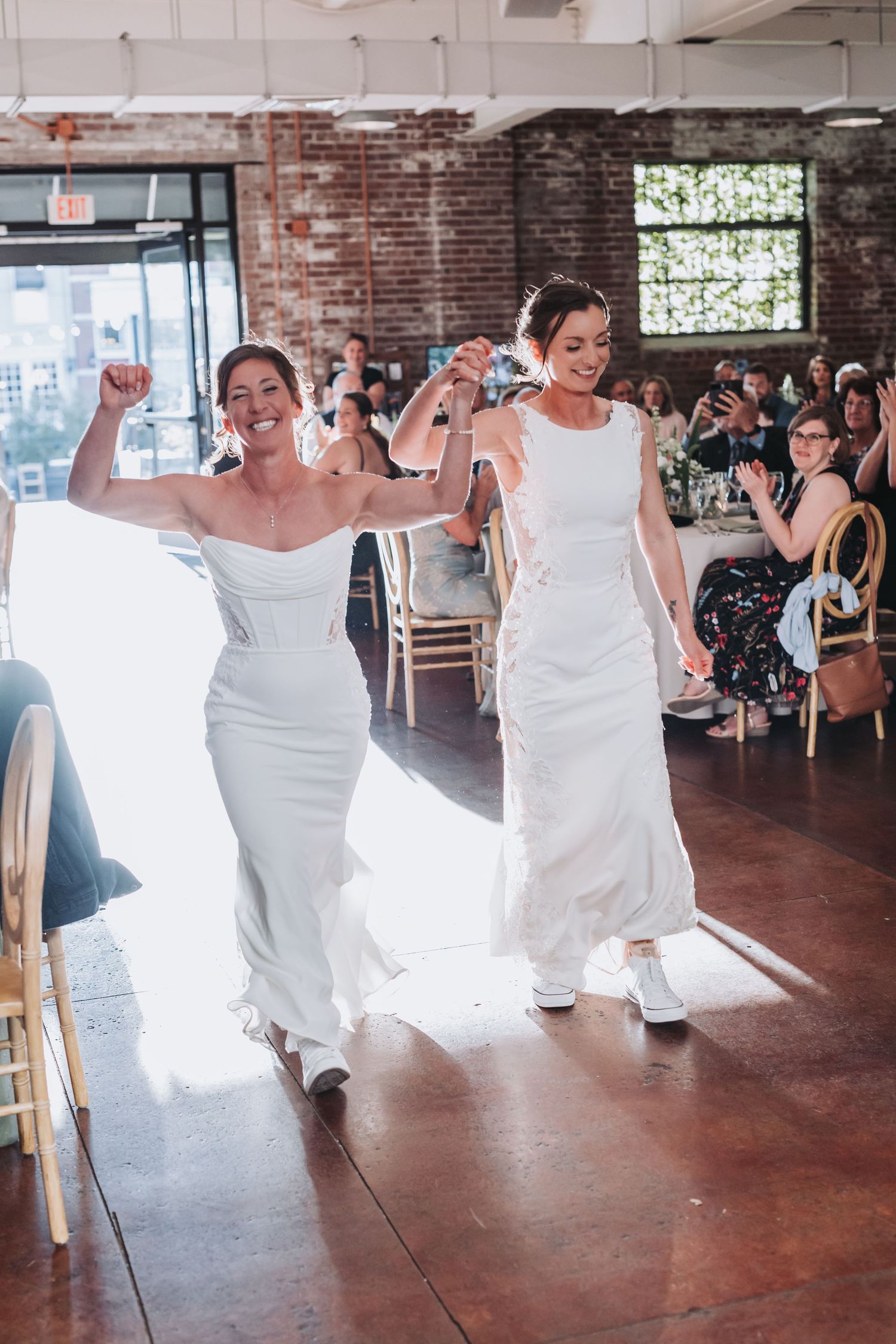 Two brides in white gowns make a joyful entrance at their wedding reception in a rustic brick venue.