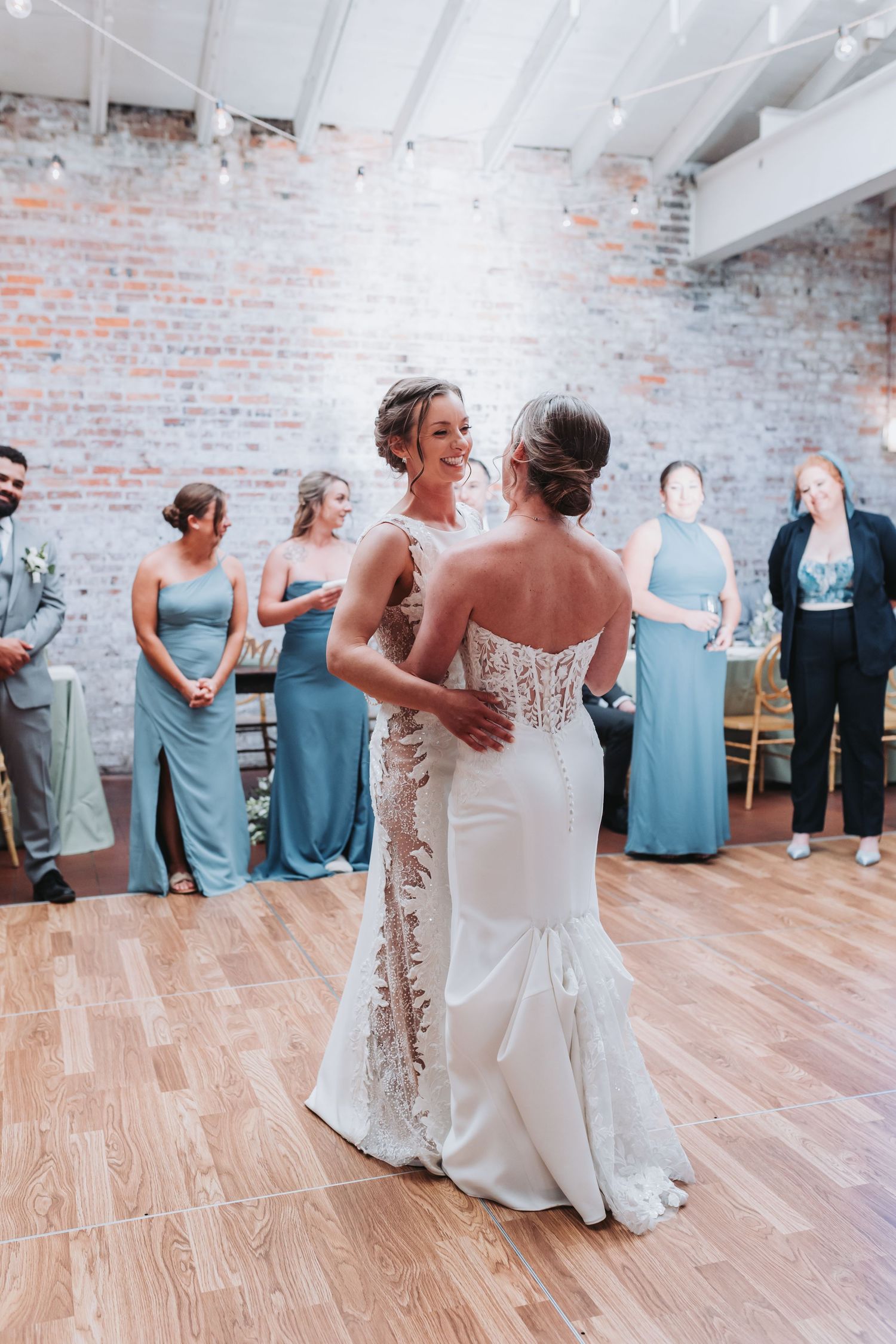 Two brides sharing first dance in white gowns surrounded by bridesmaids in blue dresses at wedding reception.