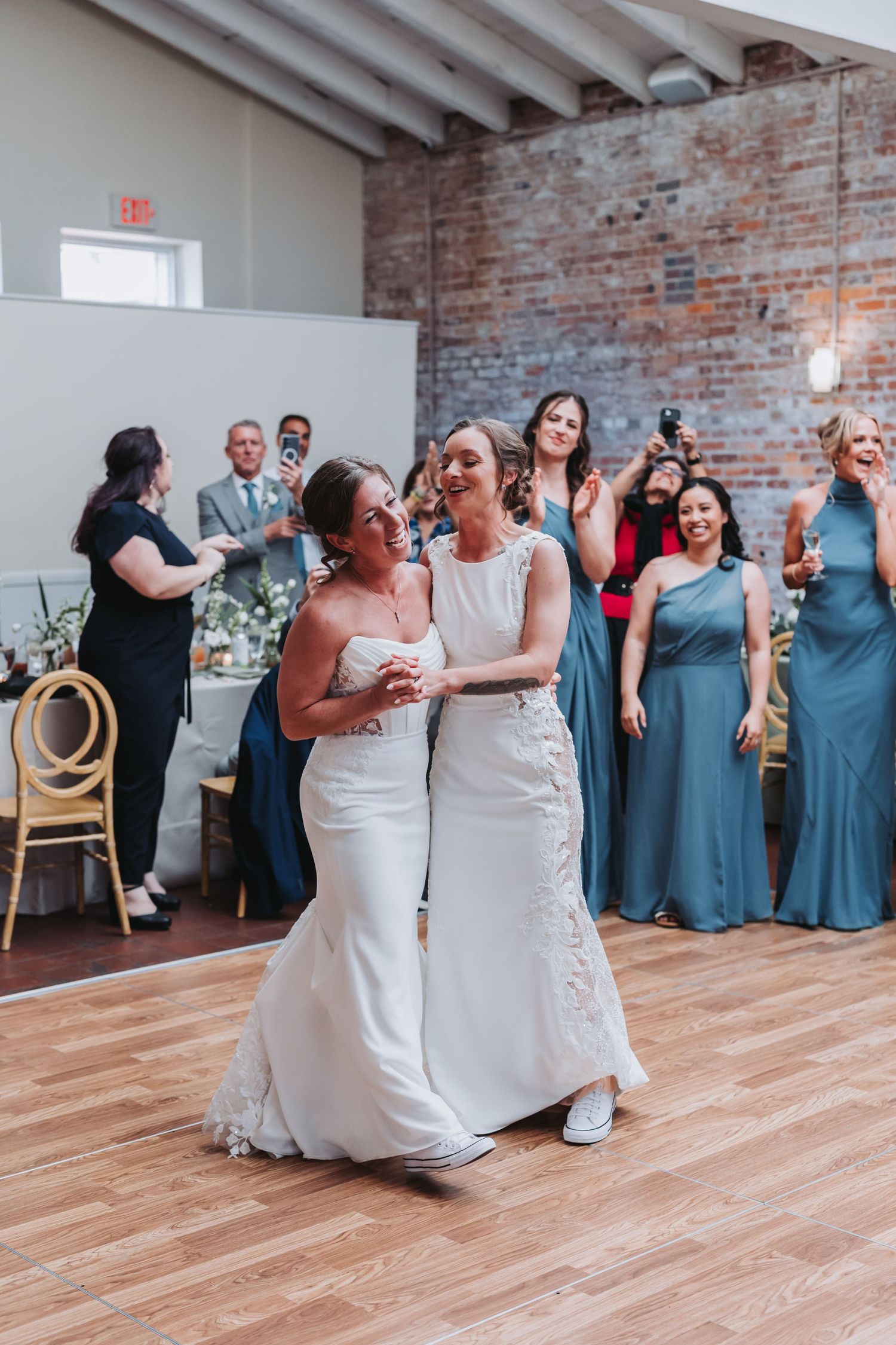 Two brides in white gowns share first dance surrounded by bridesmaids in blue dresses at wedding reception.