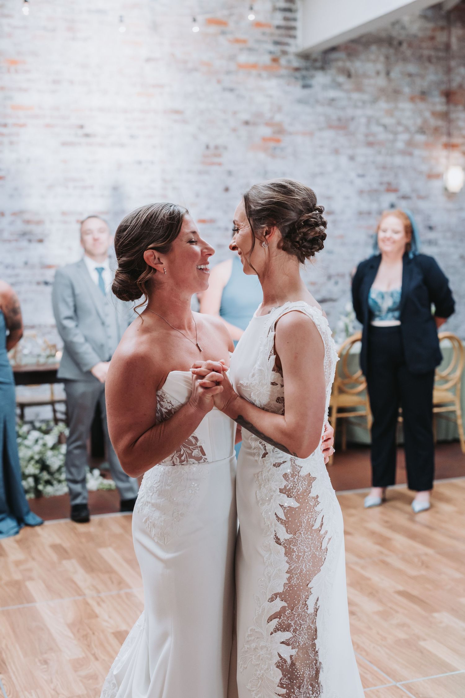 Two brides sharing their first dance in elegant wedding gowns at an indoor wedding reception.