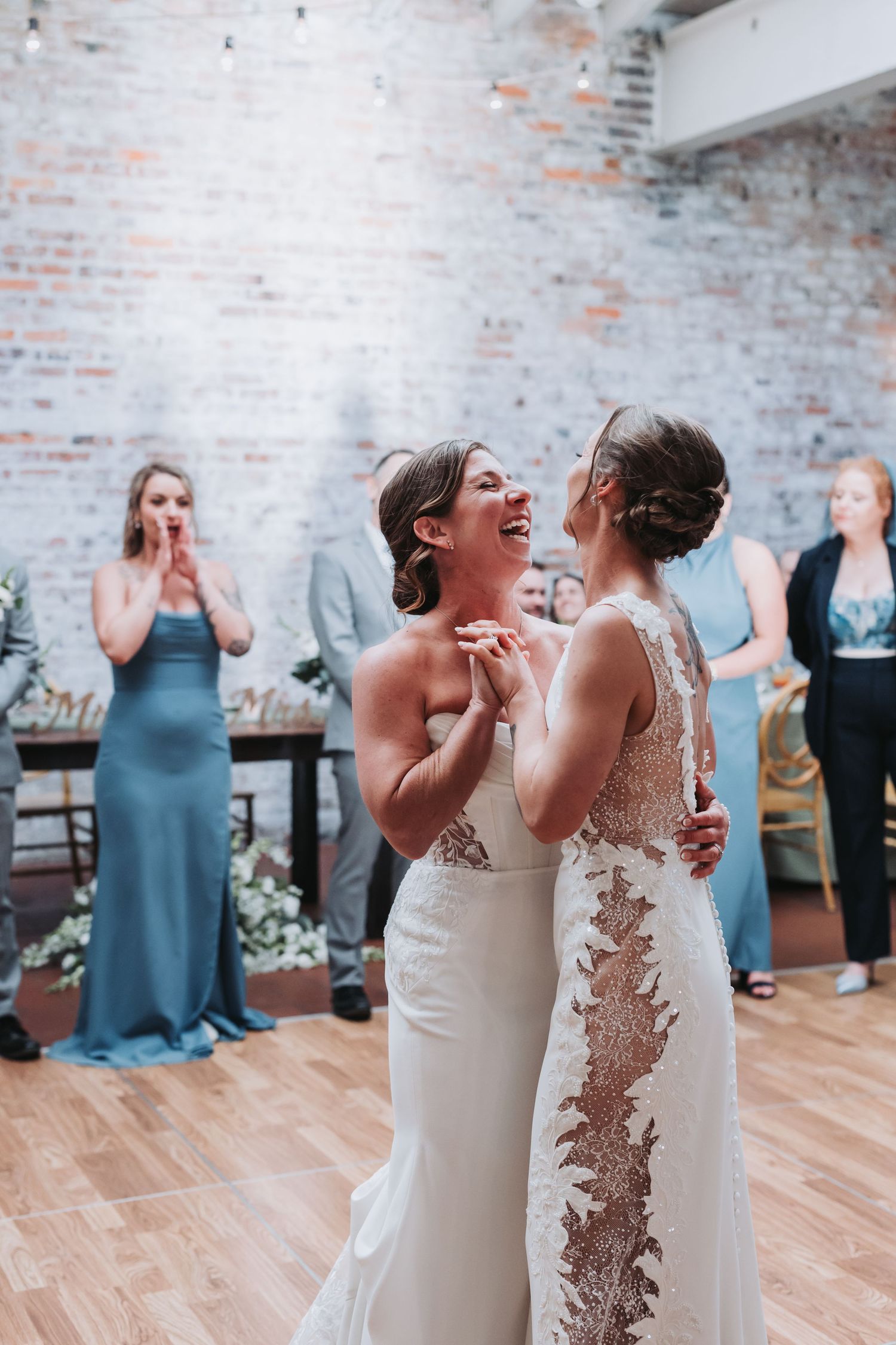 Two brides laugh joyfully during first dance at wedding reception in rustic brick wall venue.